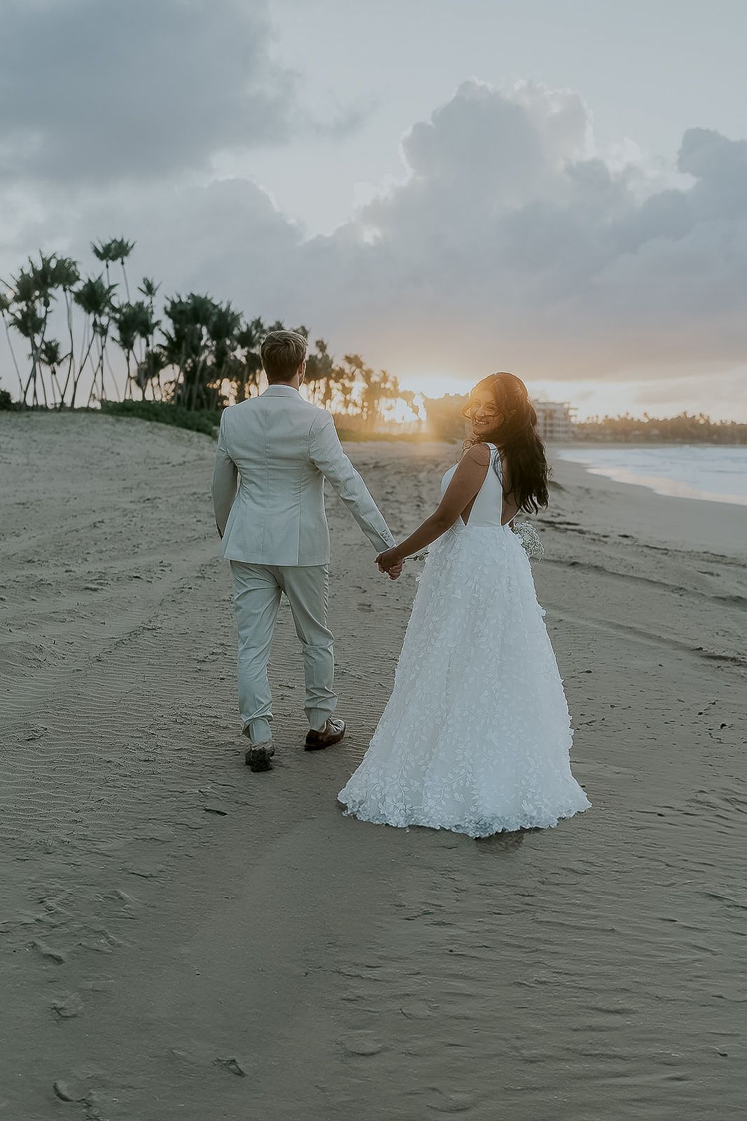 Bride and groom holding hands walking on sandy beach at sunset with palm trees in background.