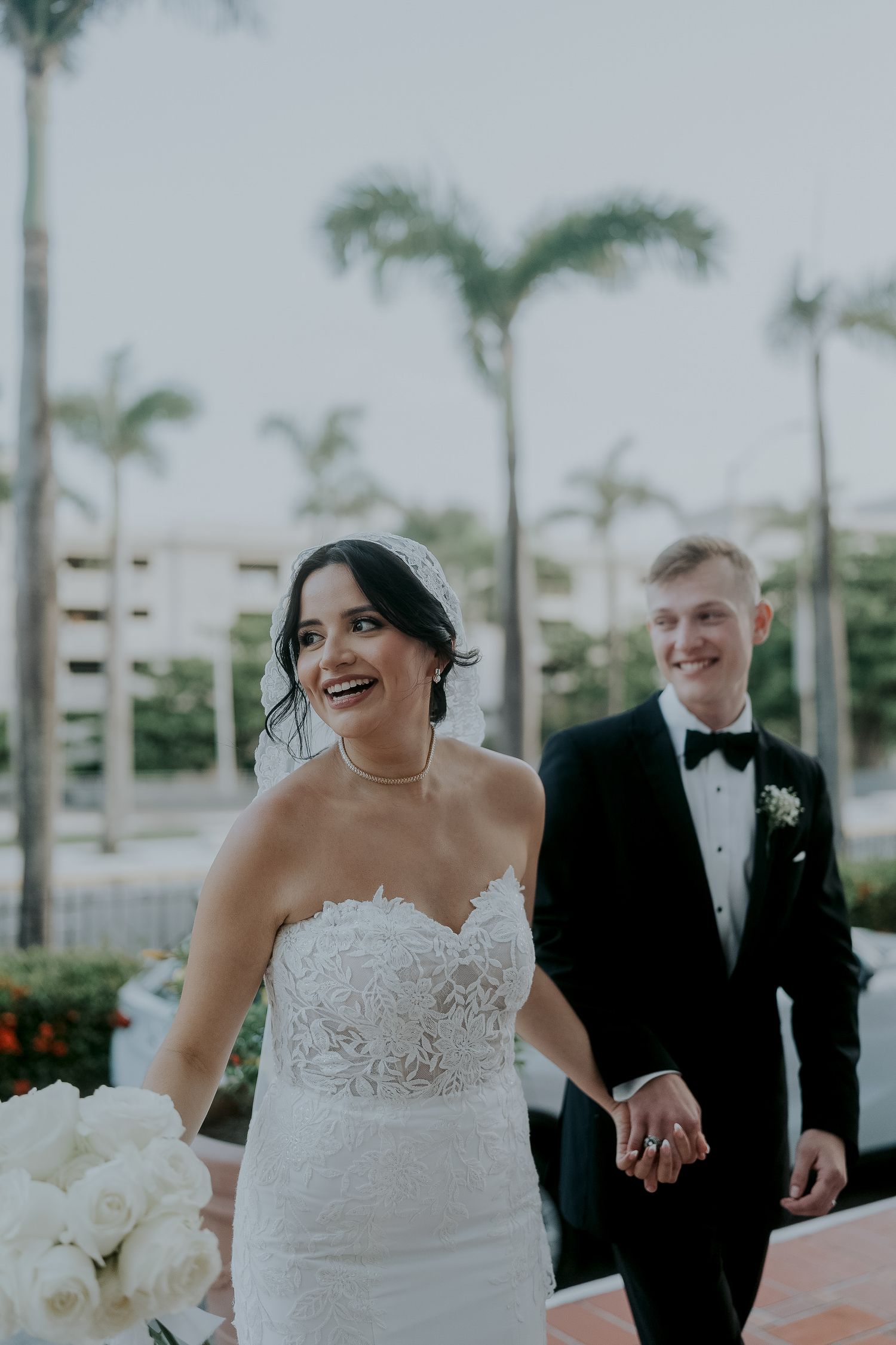 Smiling bride in strapless lace gown holding white roses with groom in black tuxedo outdoors.