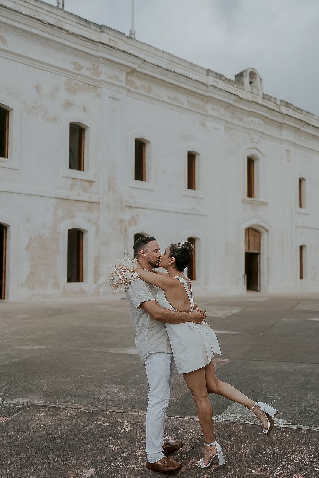 Couple sharing a romantic kiss outside a historic white stone building, woman lifting her leg holding a blush bouquet.
