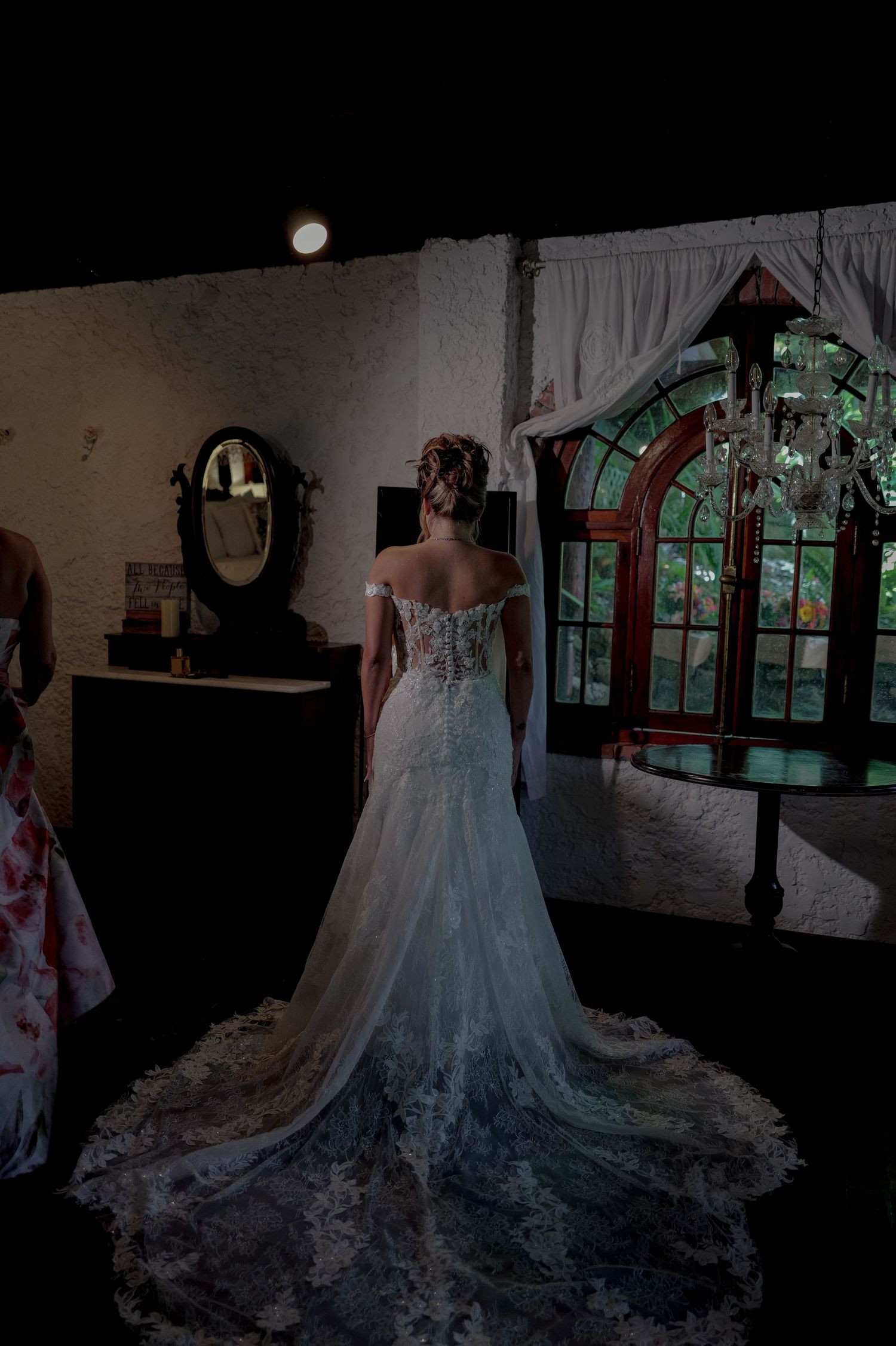 Bride in elegant lace off-shoulder wedding gown with long train standing before ornate stained glass window.