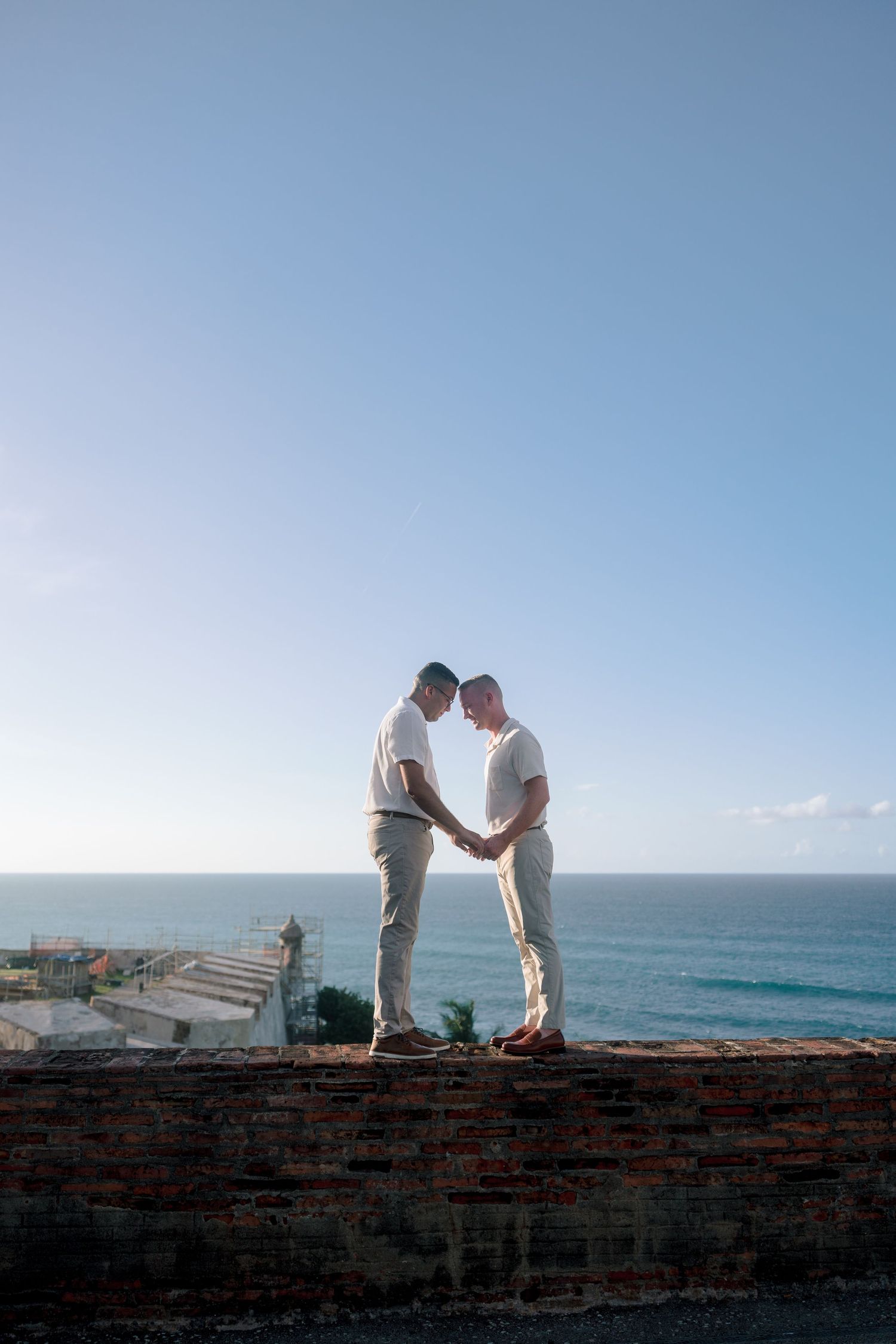 Two men holding hands and touching foreheads on a brick wall with ocean and blue sky backdrop.