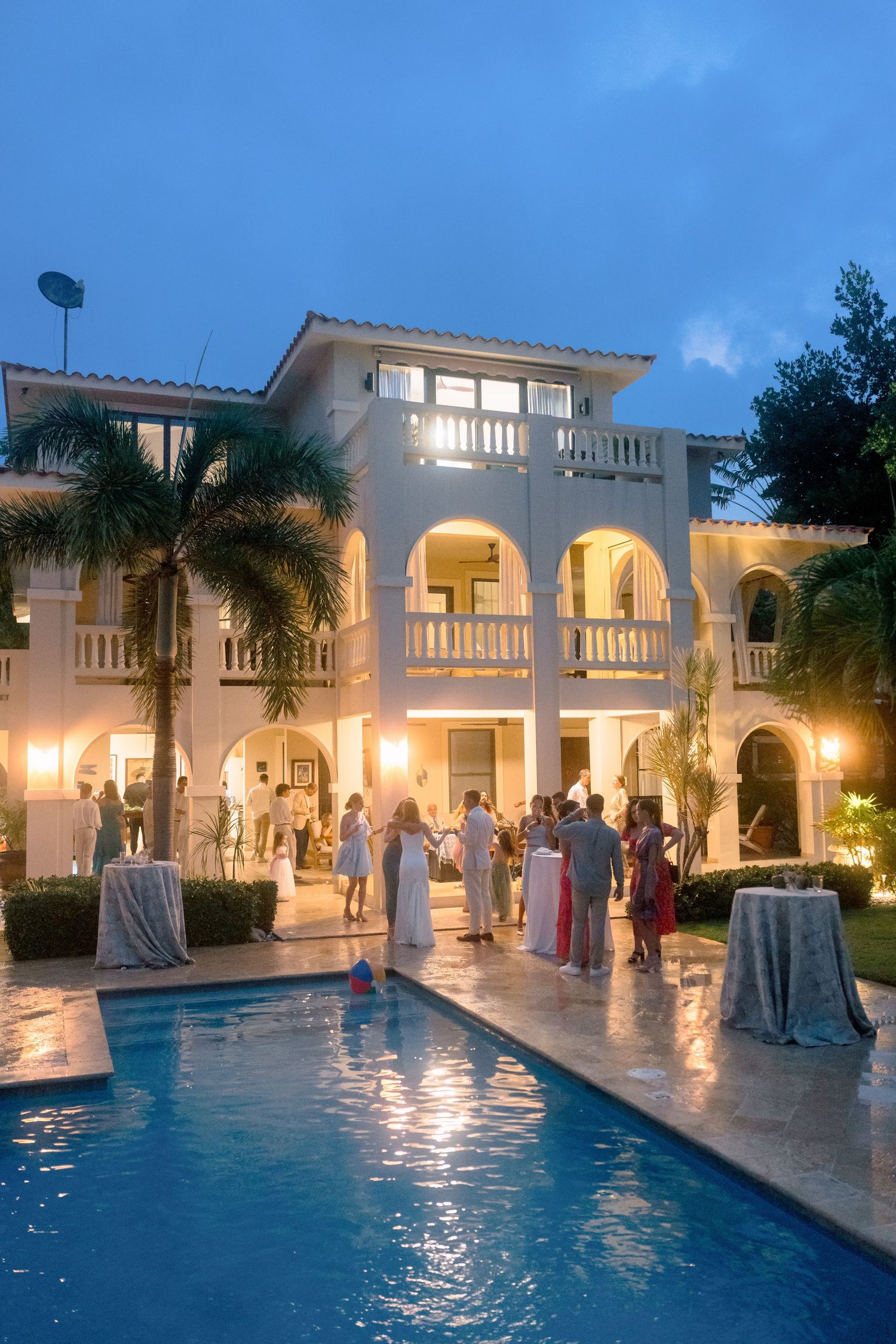 Elegant beachside villa wedding reception with pool, palm trees, and guests mingling at dusk.