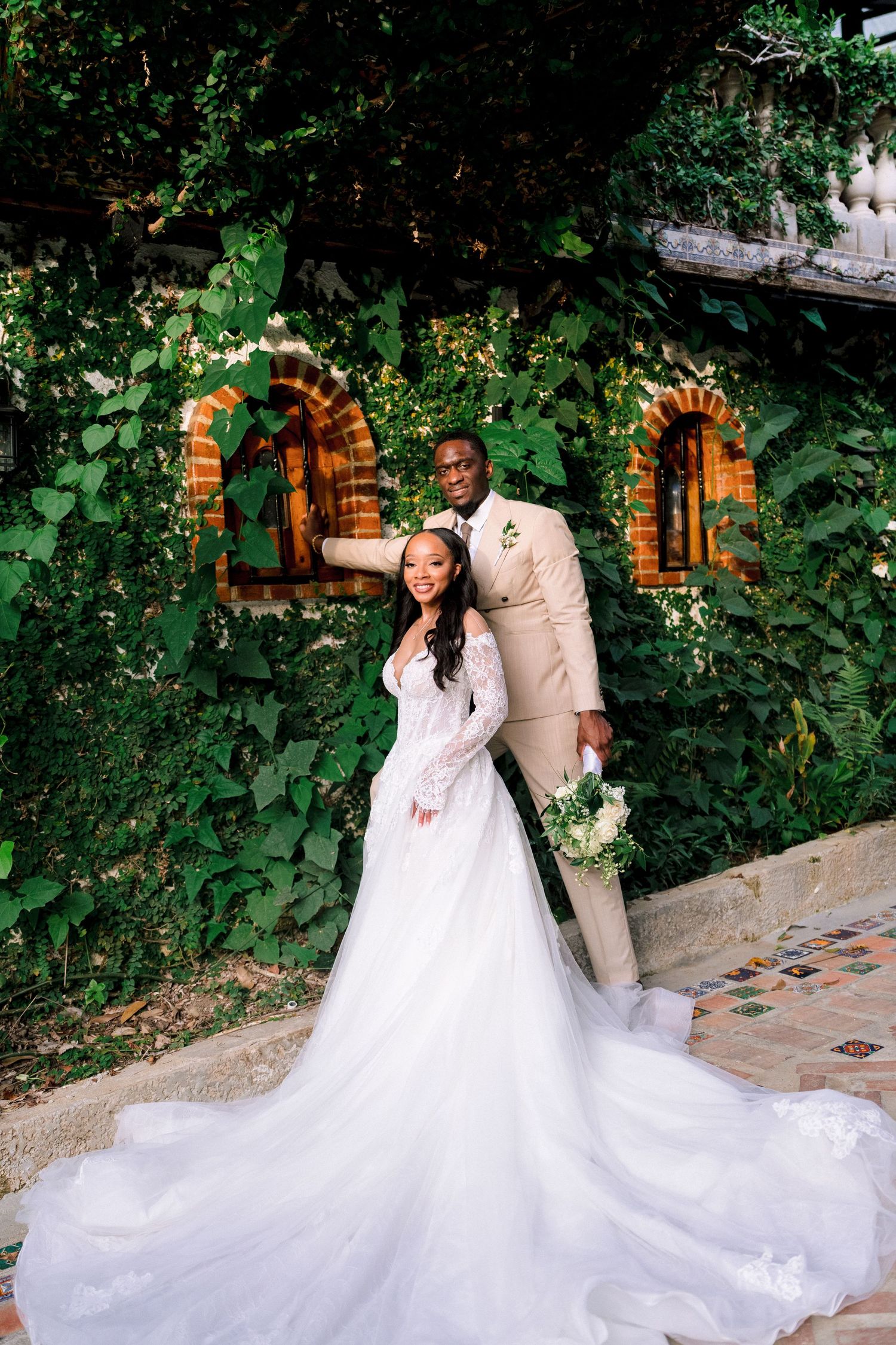 Couple poses joyfully before an ivy-covered wall, bride in a flowing lace gown holding a white bouquet.