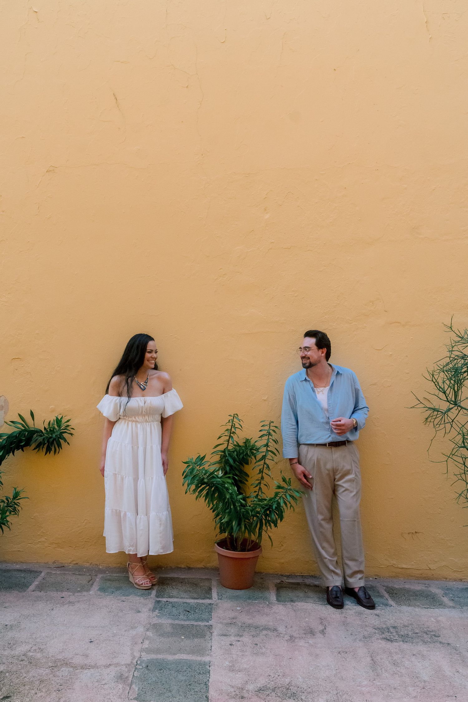 Couple in white and blue outfits standing by a vibrant yellow wall with potted plants.