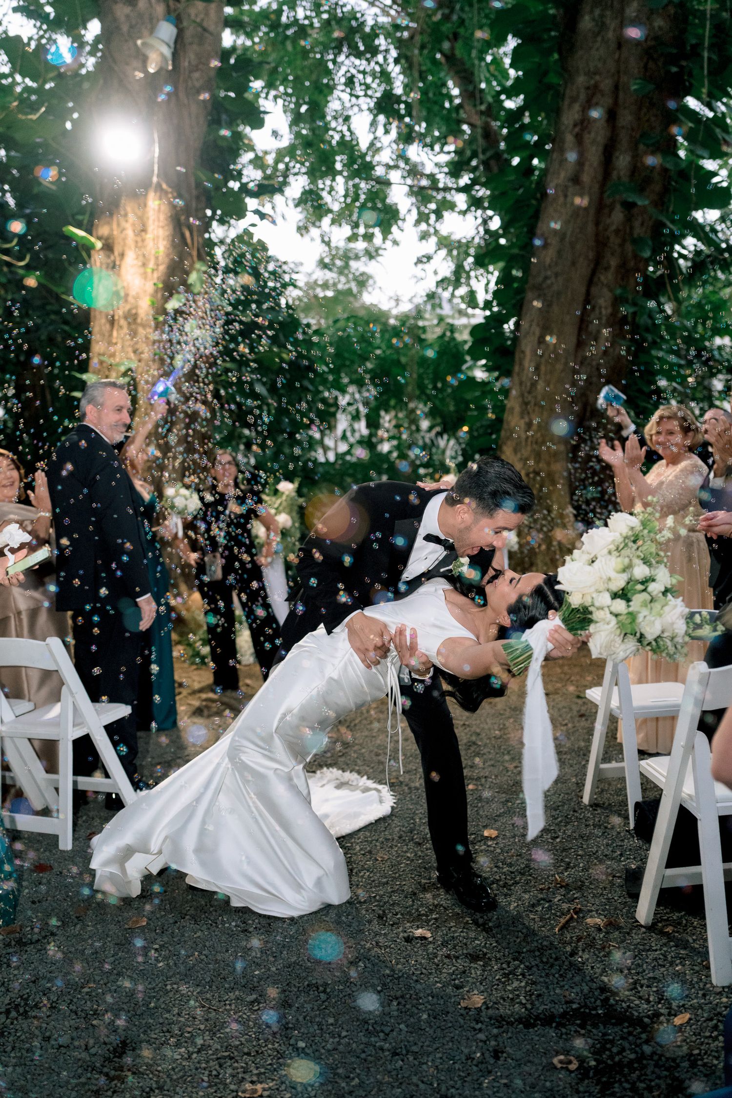 Groom dips bride for a kiss during outdoor wedding ceremony exit as guests toss confetti under tall trees.