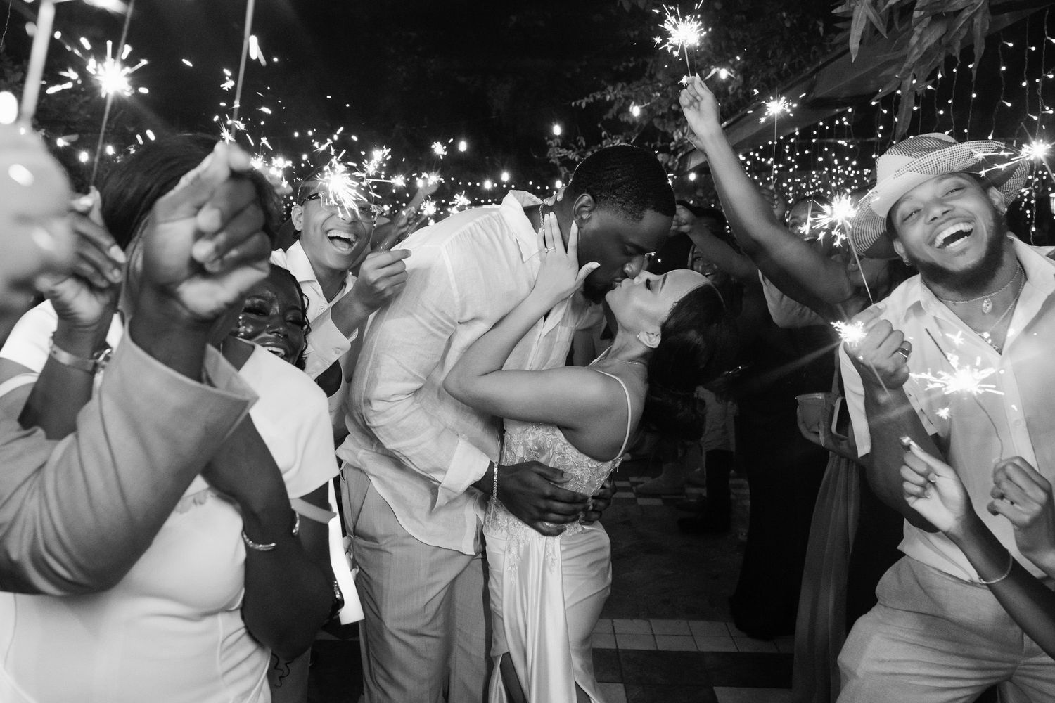 Black and white wedding photo of couple kissing surrounded by guests holding sparklers at night celebration.