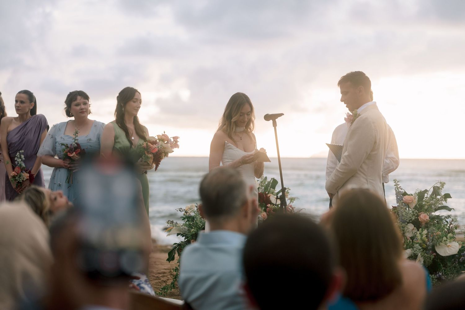 Romantic beach wedding ceremony at sunset with bride and groom exchanging vows, bridesmaids in blue dresses, ocean backdrop.
