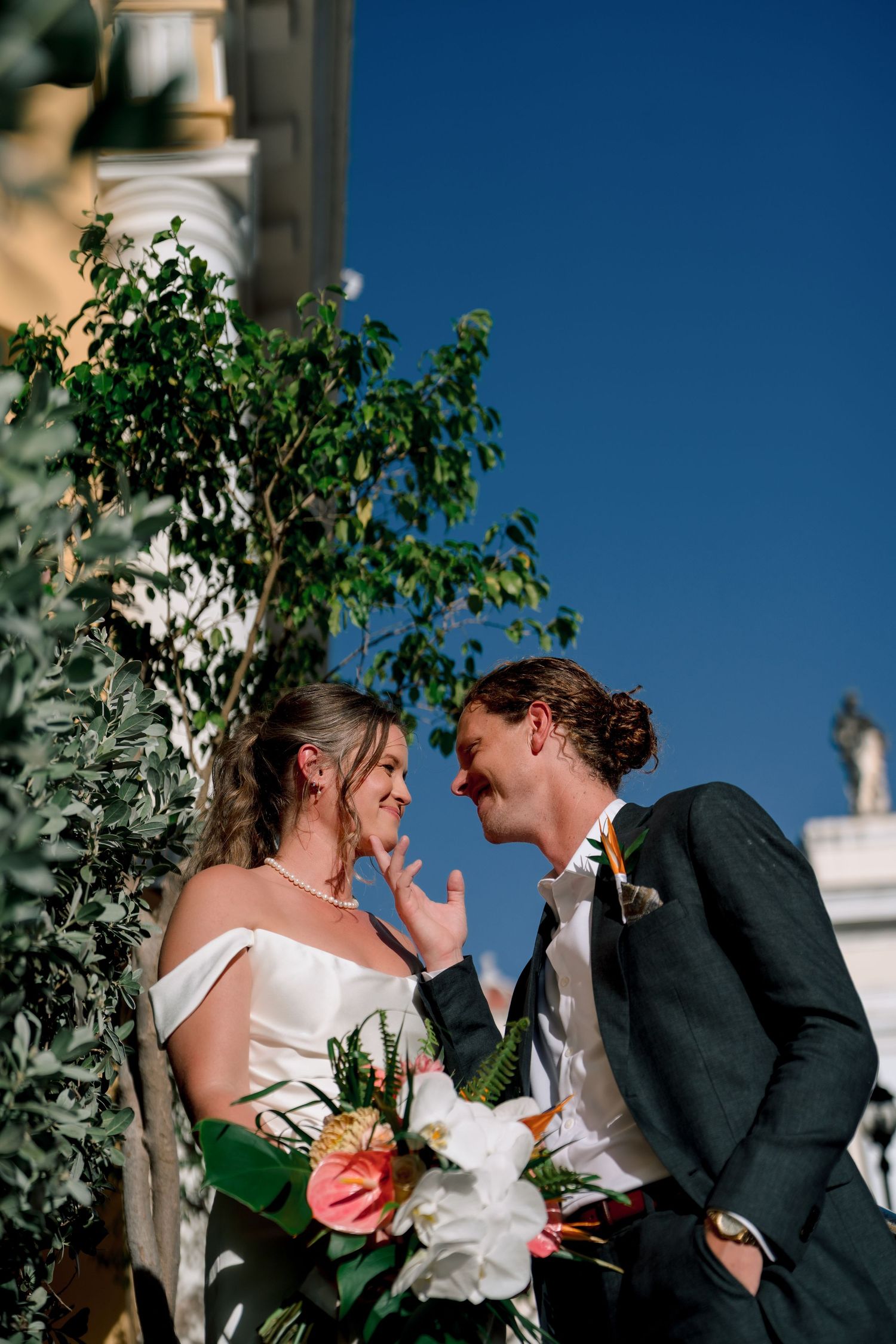 Bride and groom share a romantic moment outdoors, with lush greenery and blue sky backdrop, bride holding tropical bouquet.