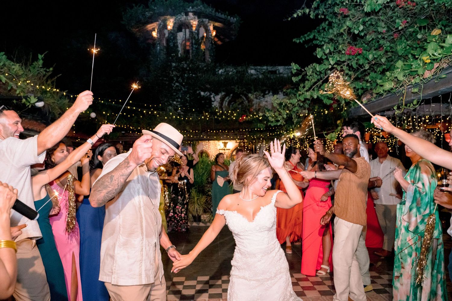 Bride and groom exit wedding reception through sparkler tunnel with guests celebrating at night outdoor venue.