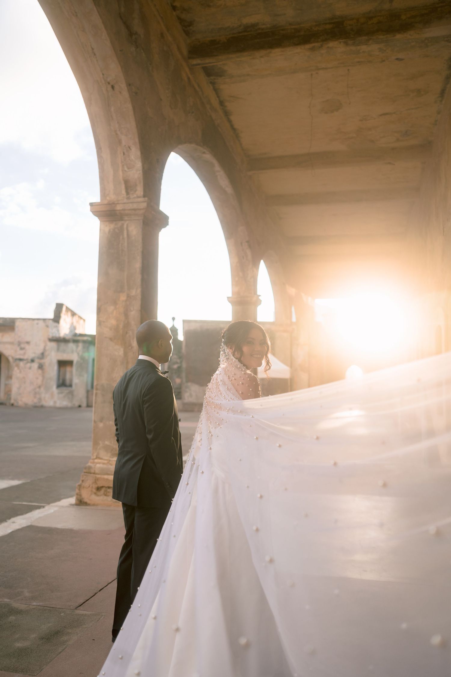 Wedding couple stands beneath stone arch as golden sunset light illuminates bride's flowing pearl veil outdoors.
