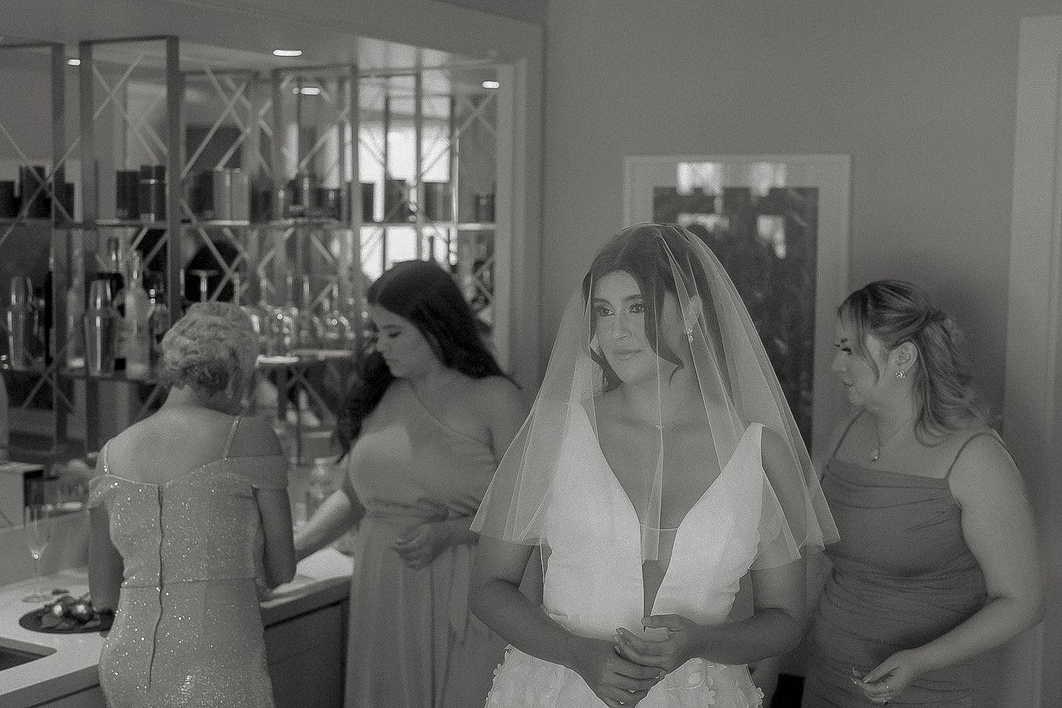 Black and white photo of a bride in a veil with bridesmaids helping her prepare for her wedding day.