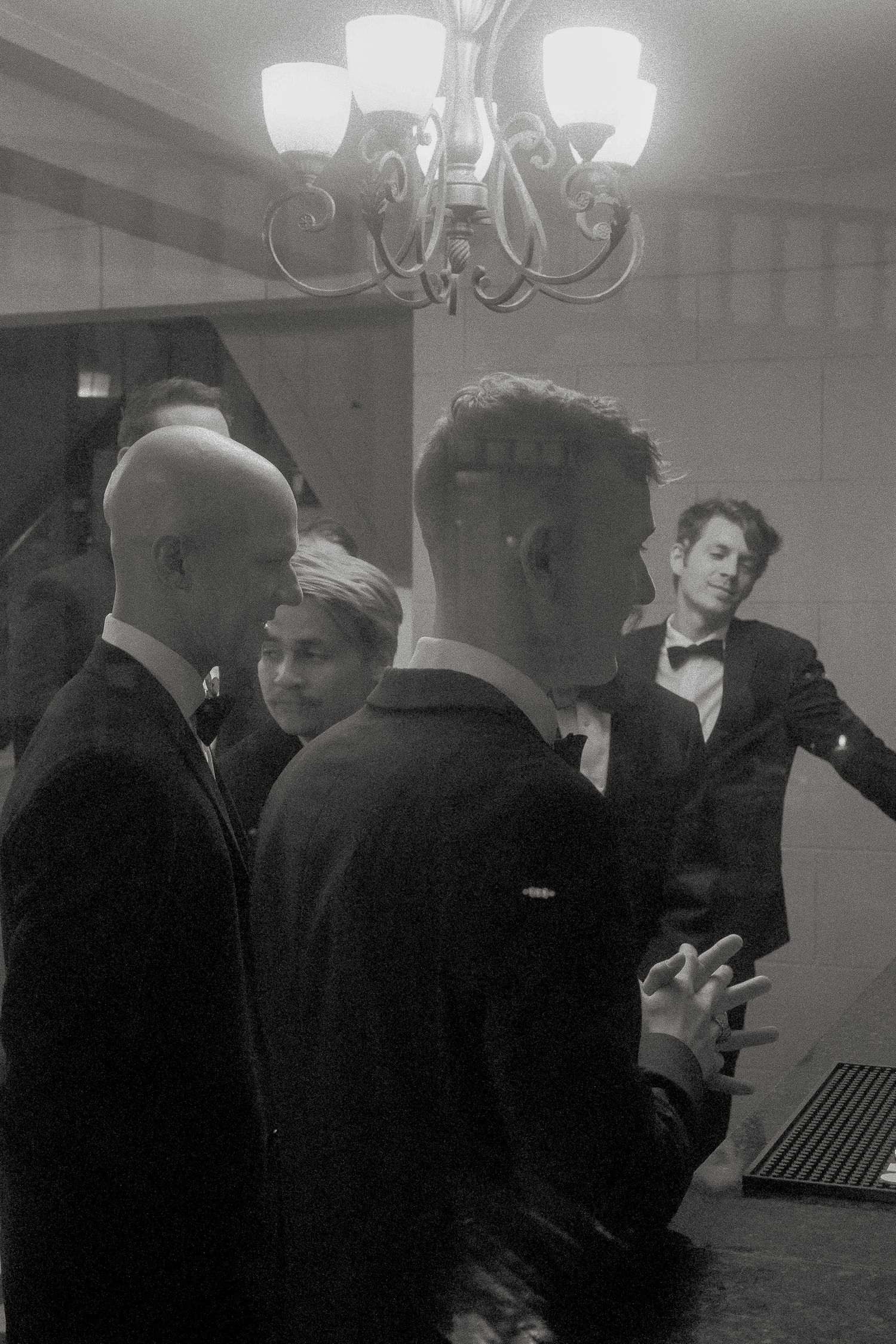 Groomsmen in tuxedos gather under chandelier in black and white wedding photo.