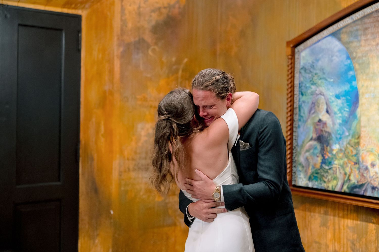 Bride and groom share an emotional first look hug in a warmly lit venue with a colorful painting on the wall.