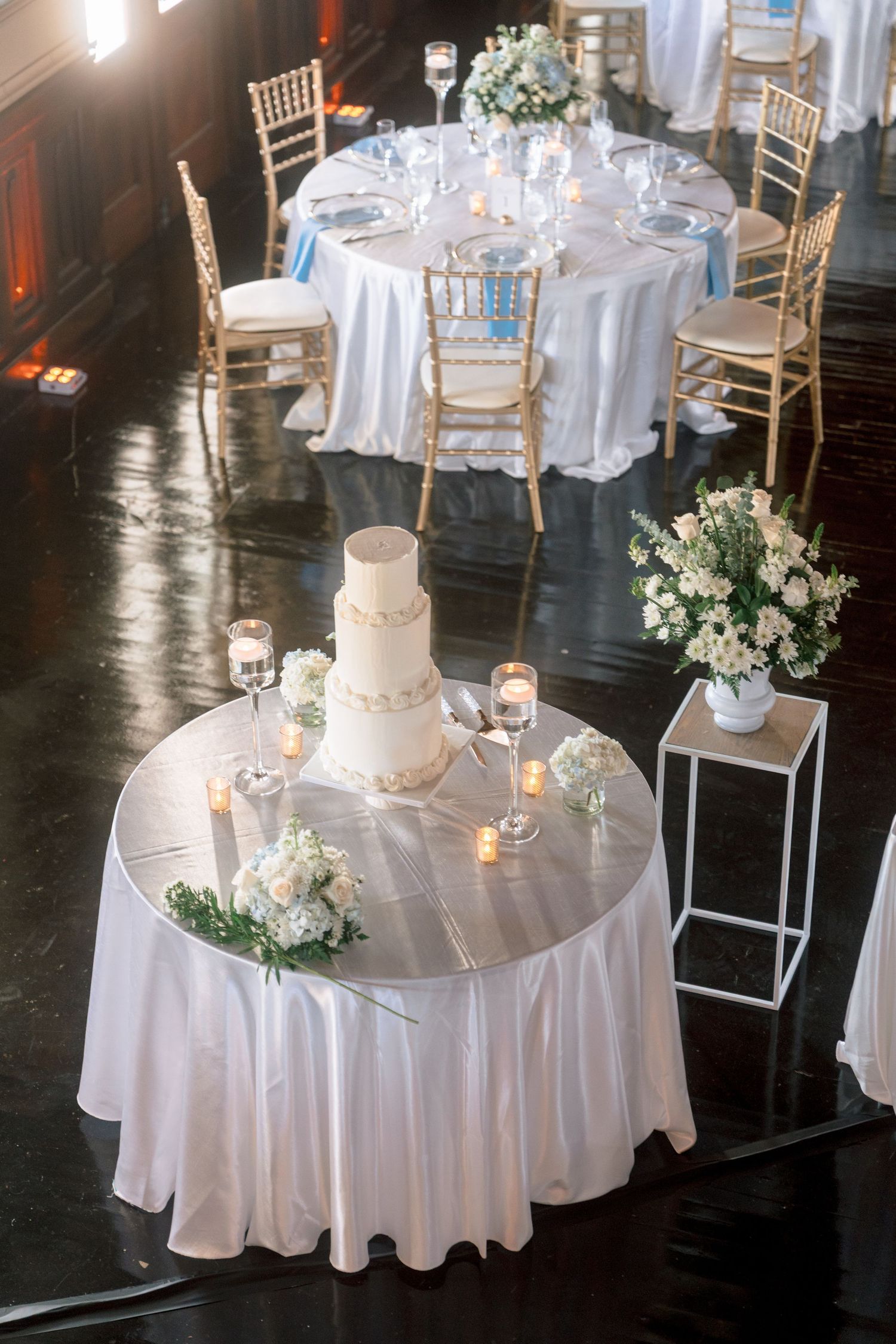 Elegant wedding reception with a tiered white cake on a draped table, candles, floral arrangements, and gold Chiavari chairs.