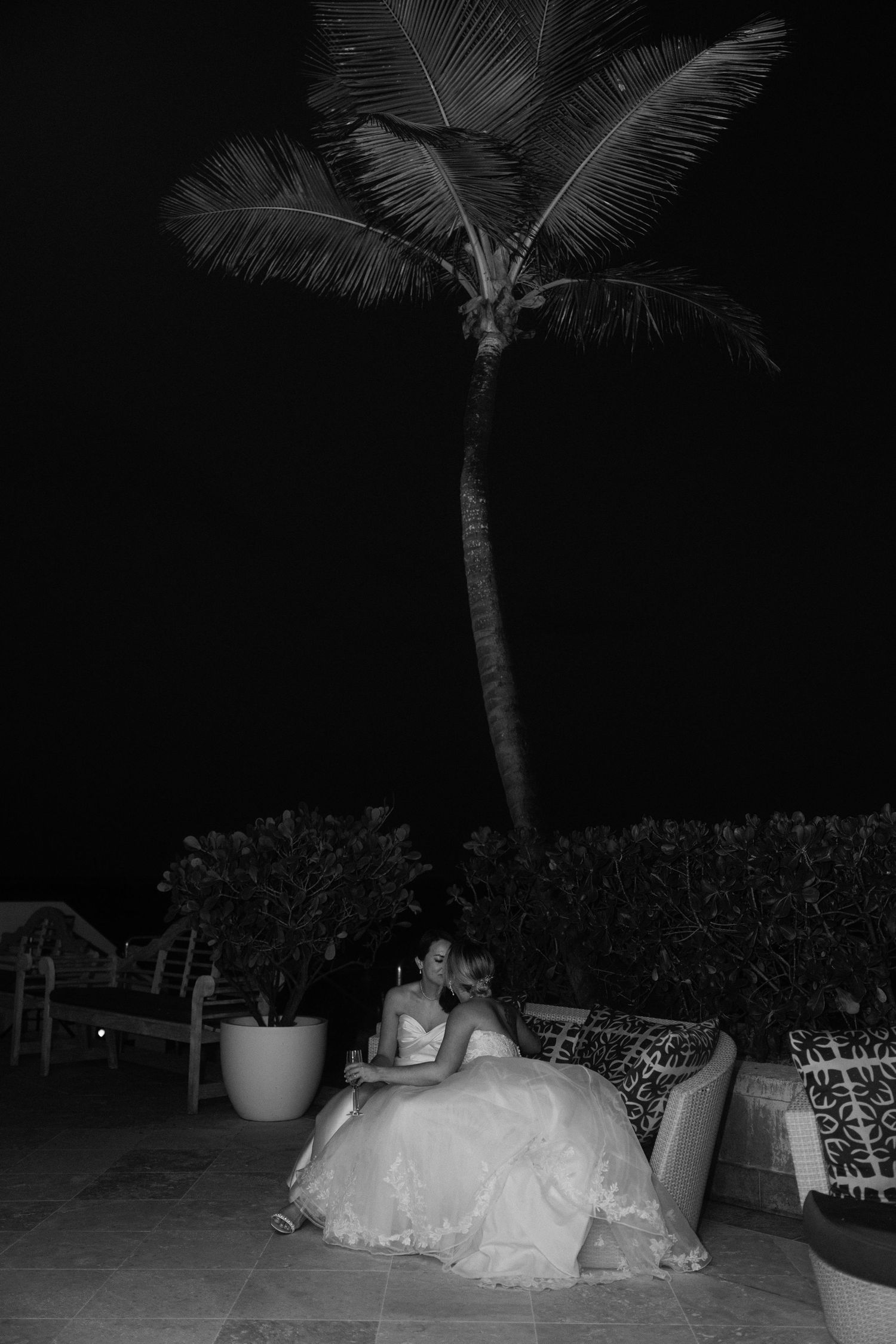 Bride in white gown sitting outdoors at night under a palm tree in a black and white wedding photo.