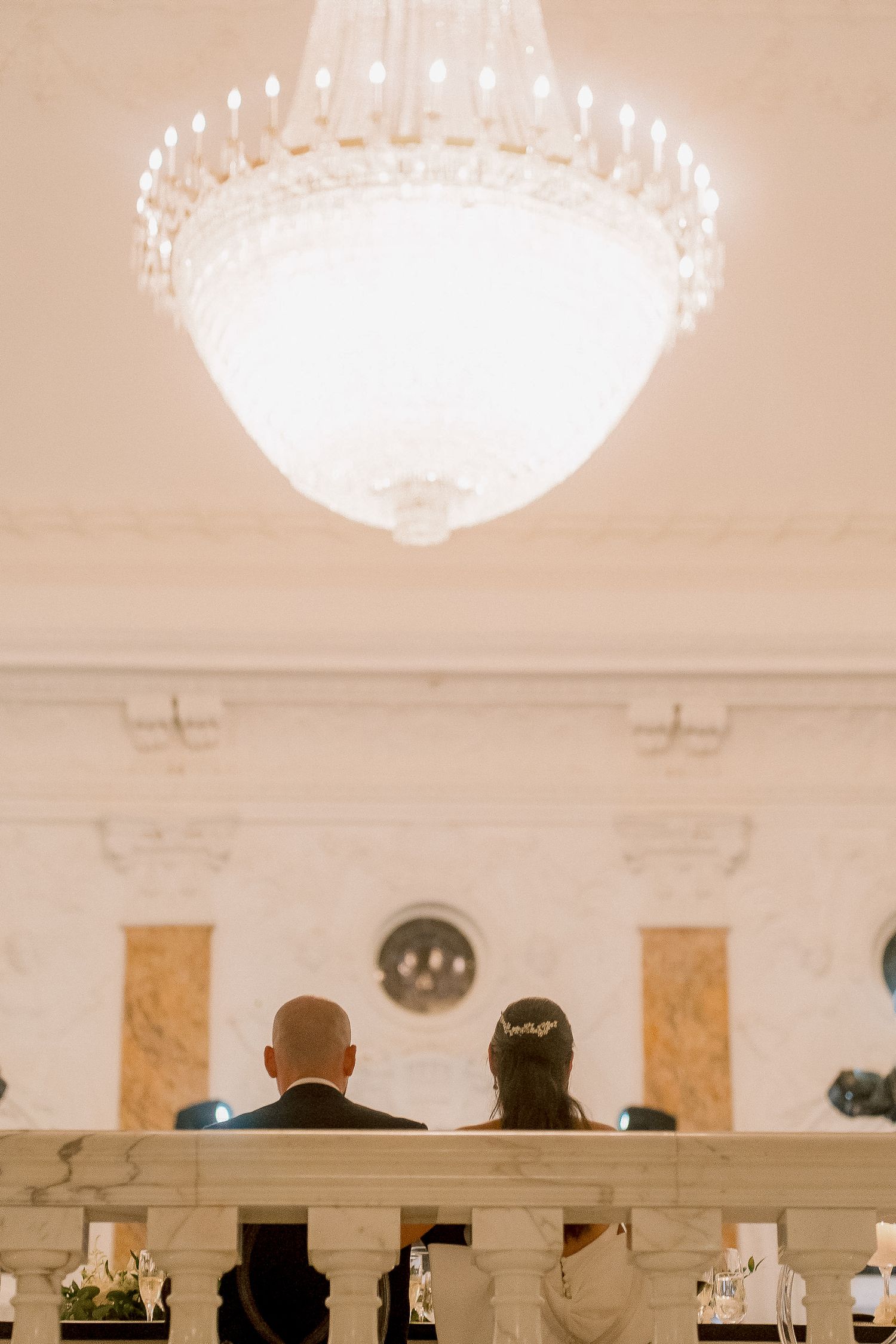 Bride and groom seated at altar beneath grand crystal chandelier in elegant white wedding venue.