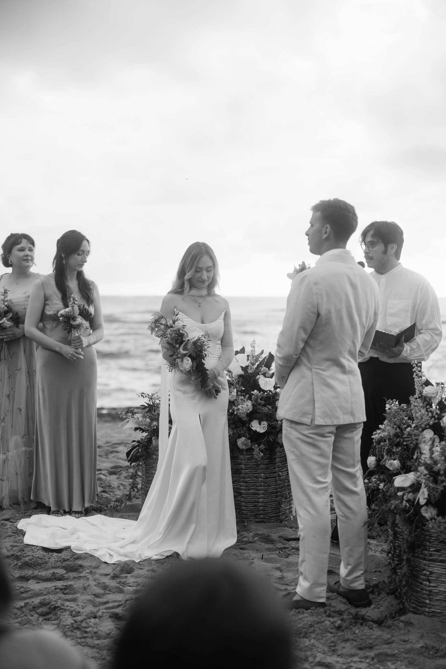 Black and white beach wedding ceremony with bride holding bouquet facing groom, bridesmaids and officiant beside them.