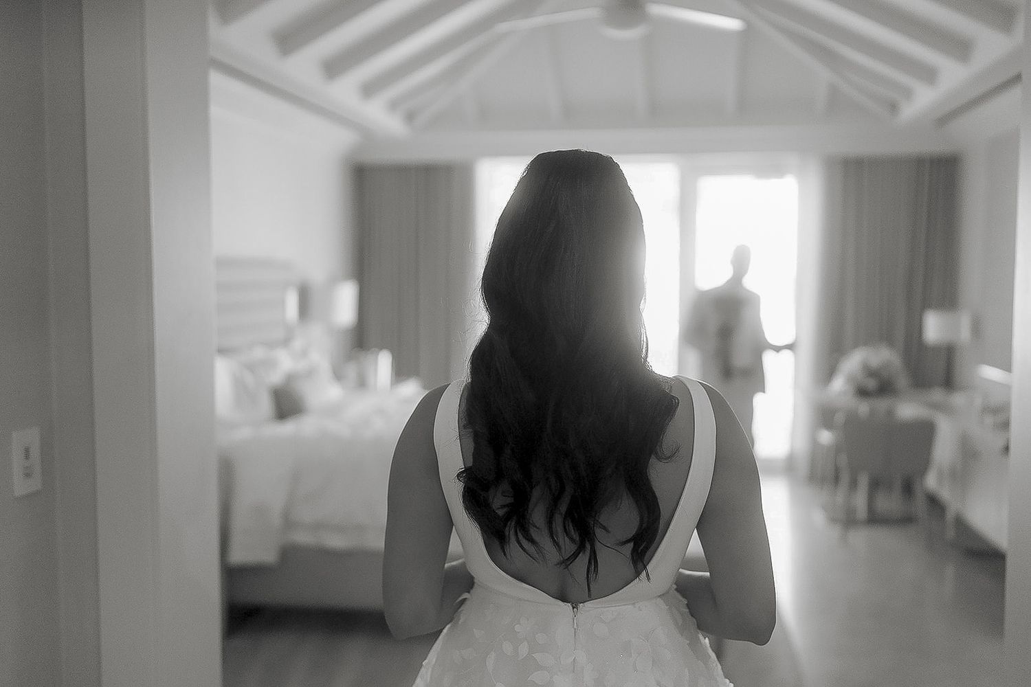 Black and white photo of a bride in a wedding dress standing in a hotel room, viewed from behind.