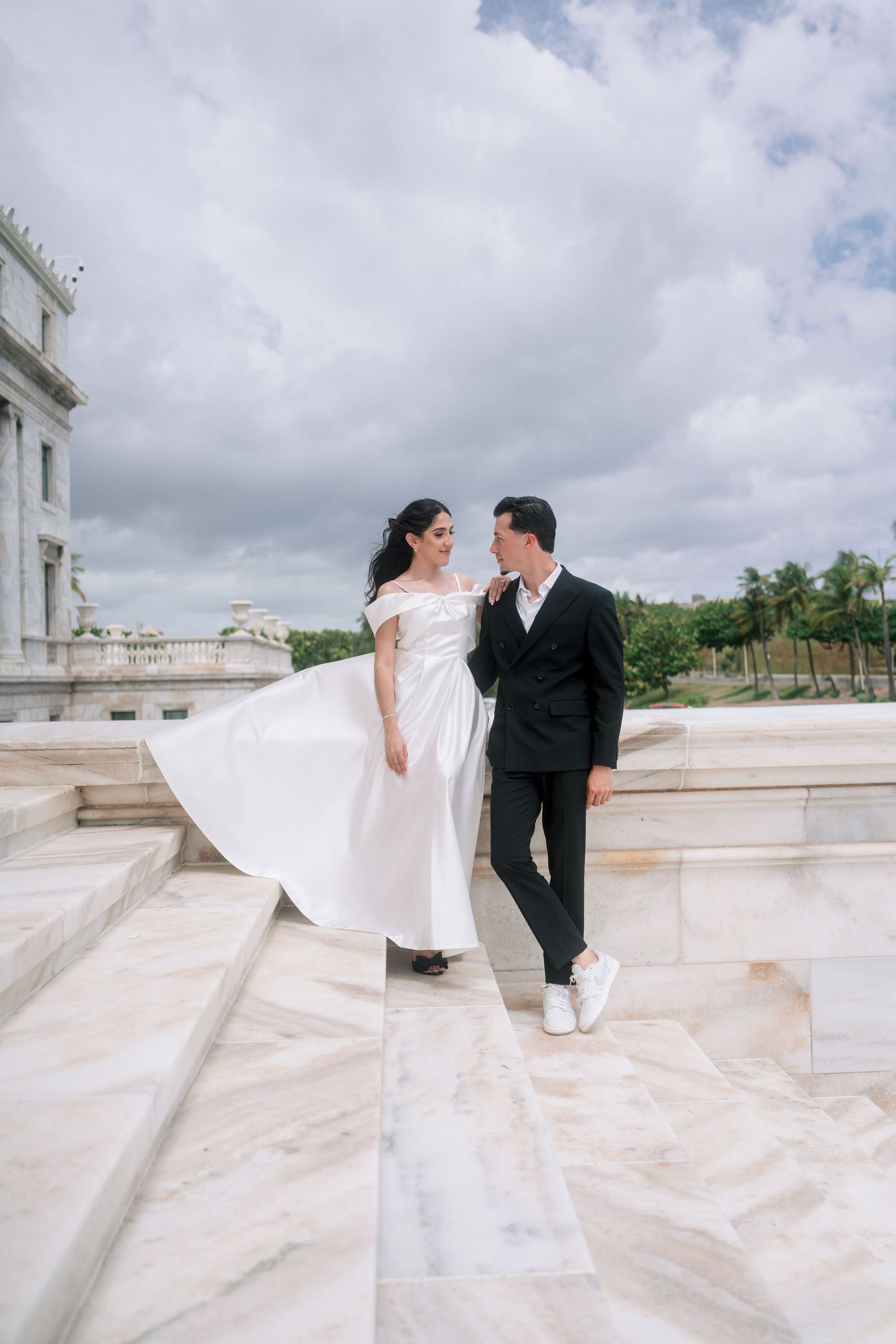 Bride in white off-shoulder gown and groom in black suit share a romantic moment on marble steps outdoors.