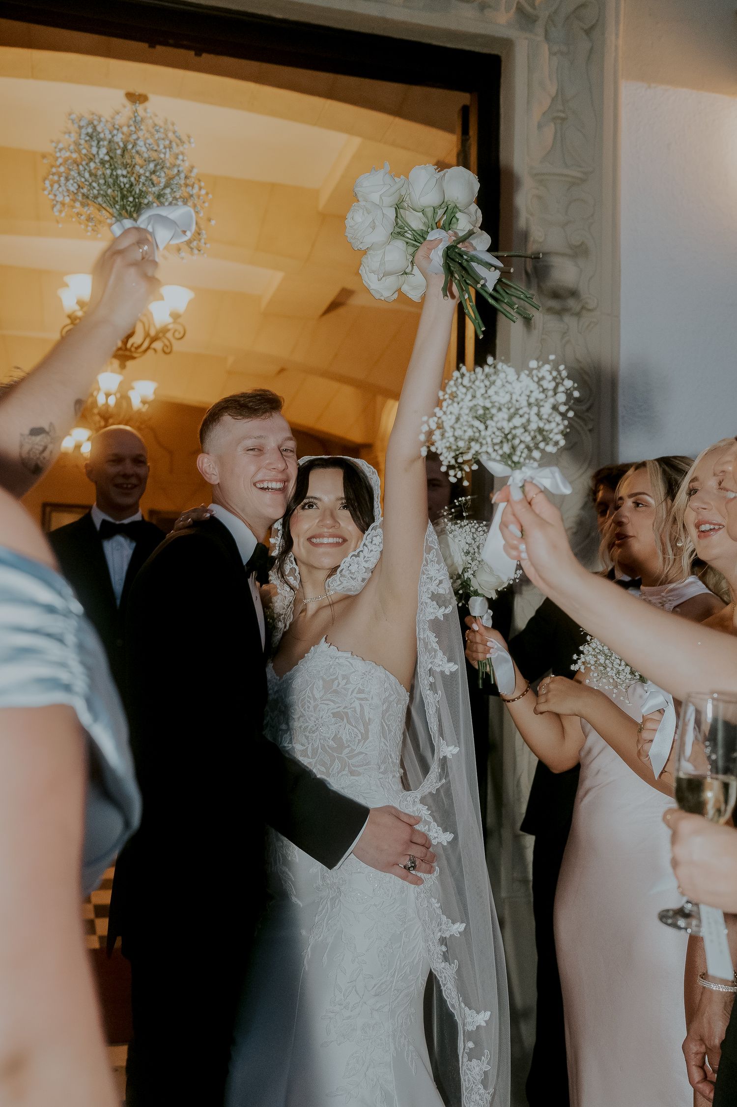 Joyful bride and groom exit ceremony raising white bouquets surrounded by cheering wedding guests.