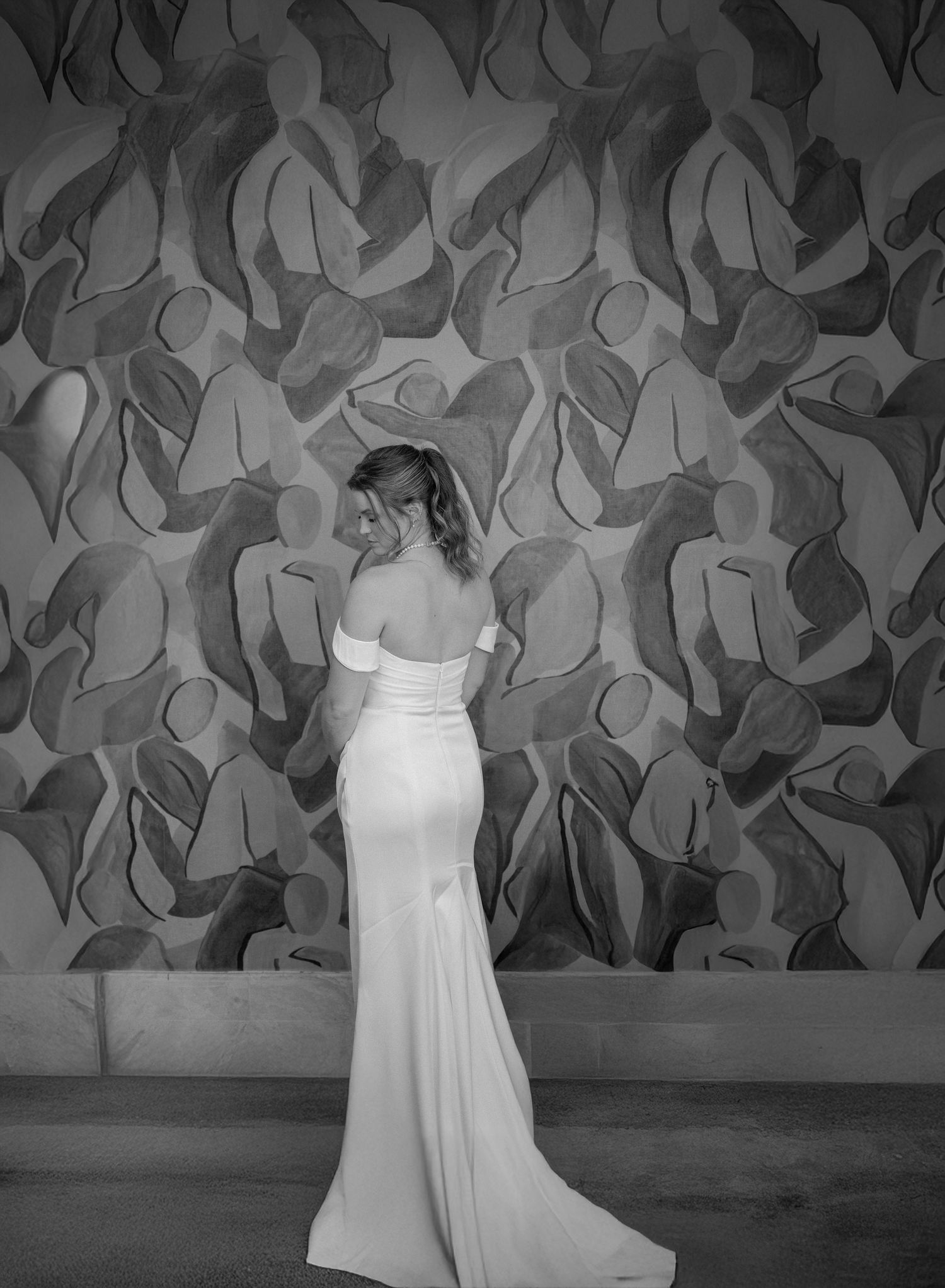 Bride in elegant off-shoulder white gown poses against abstract mural wall in black and white photo.