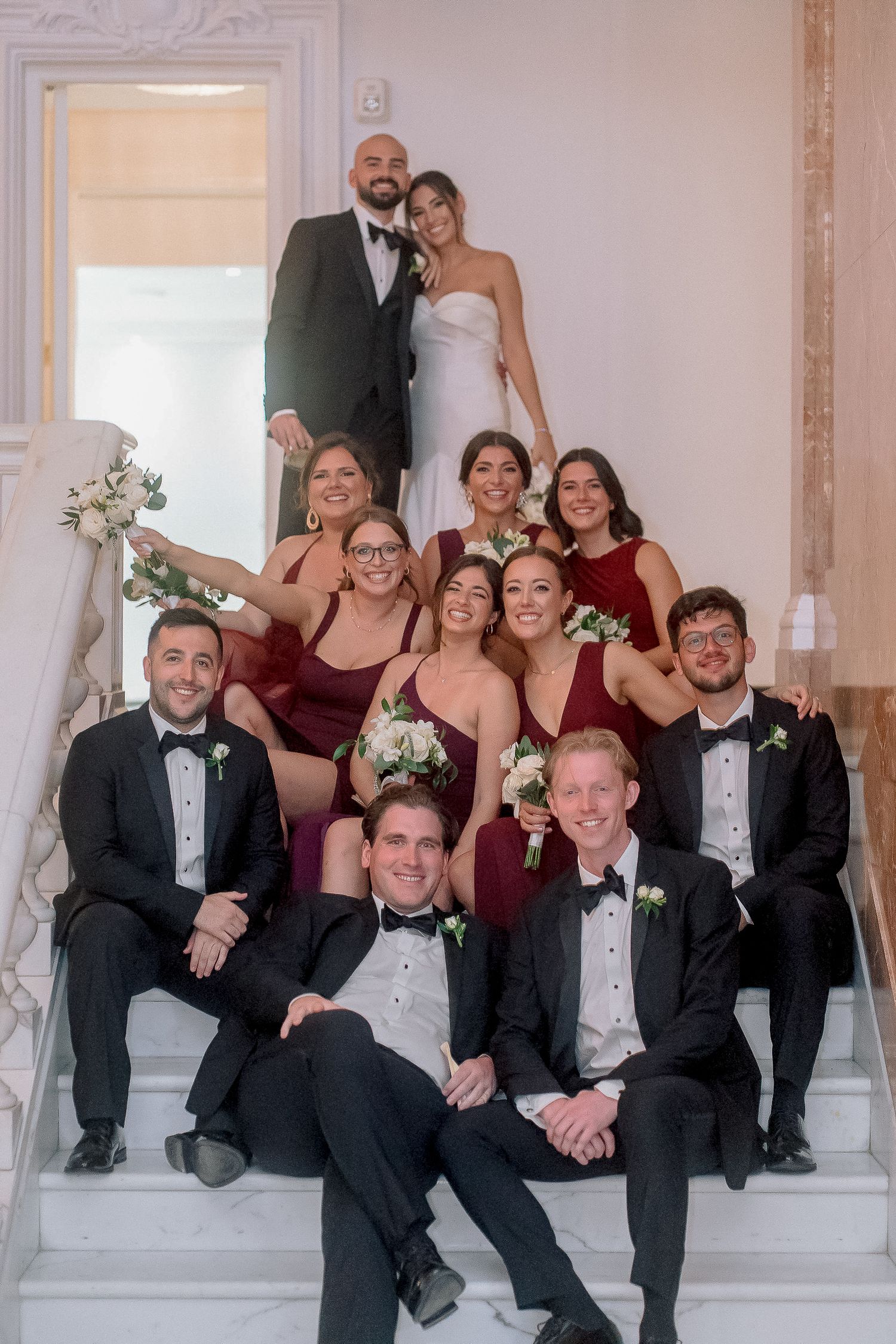 Wedding party in burgundy and black tuxedos pose on elegant marble staircase with bride and groom at top.
