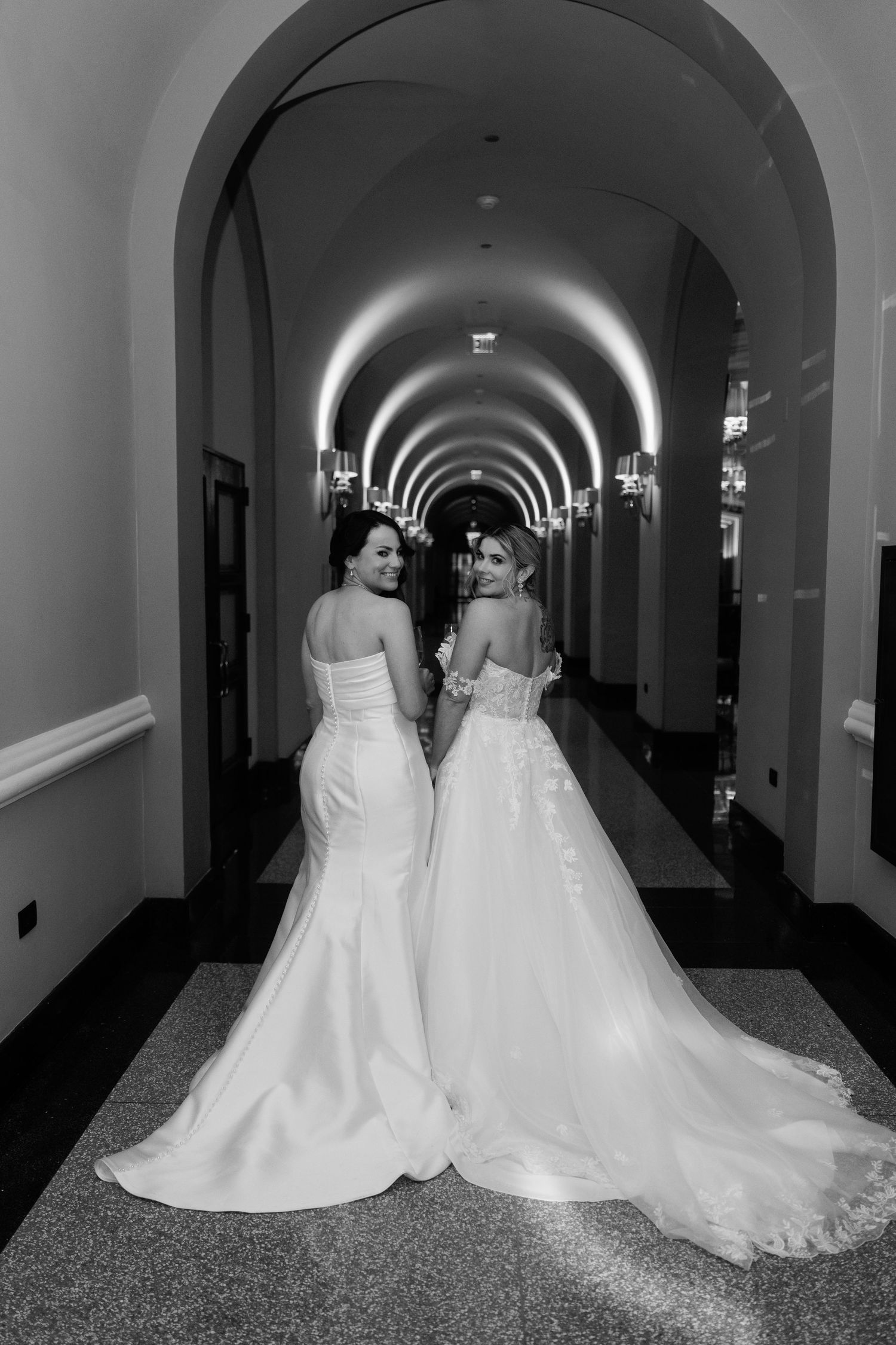 Two brides in elegant white gowns pose in a grand arched hallway, looking back in a black and white wedding photo.