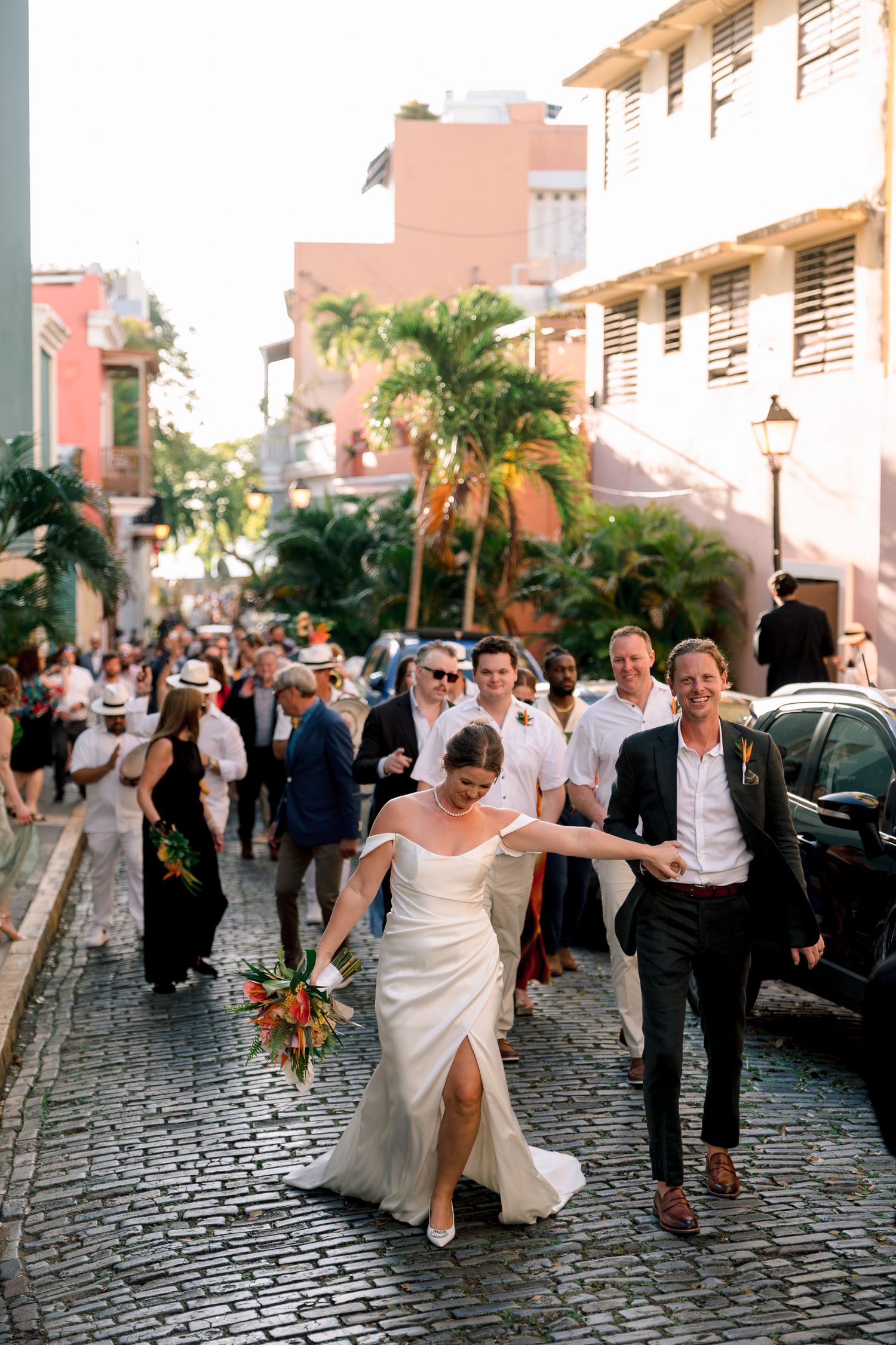 Bride in white gown walks cobblestone street with guests in tropical wedding procession in Old San Juan.