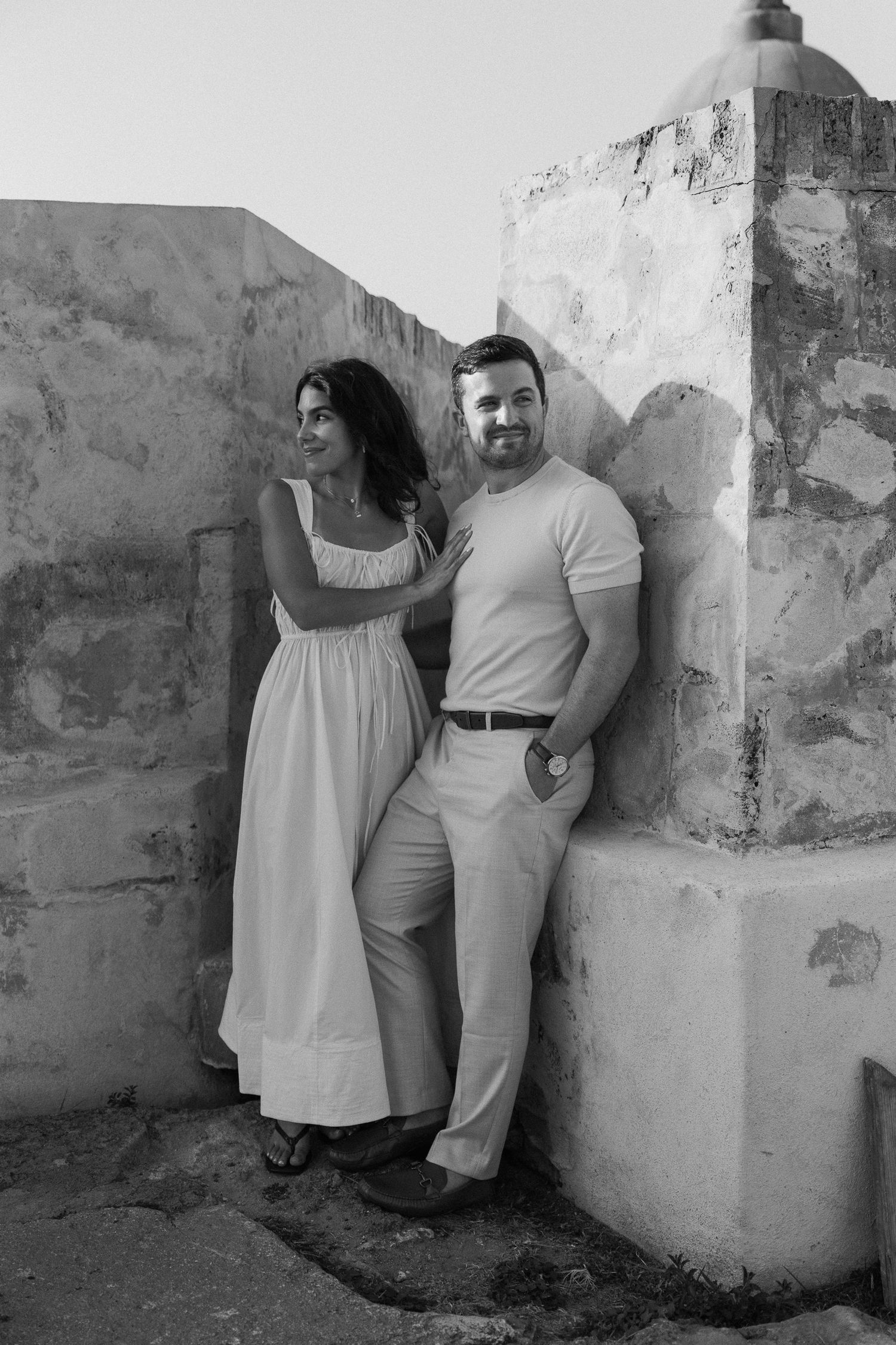 Black and white photo of a couple posing against a rustic stone wall, woman in a white dress and man in casual attire.