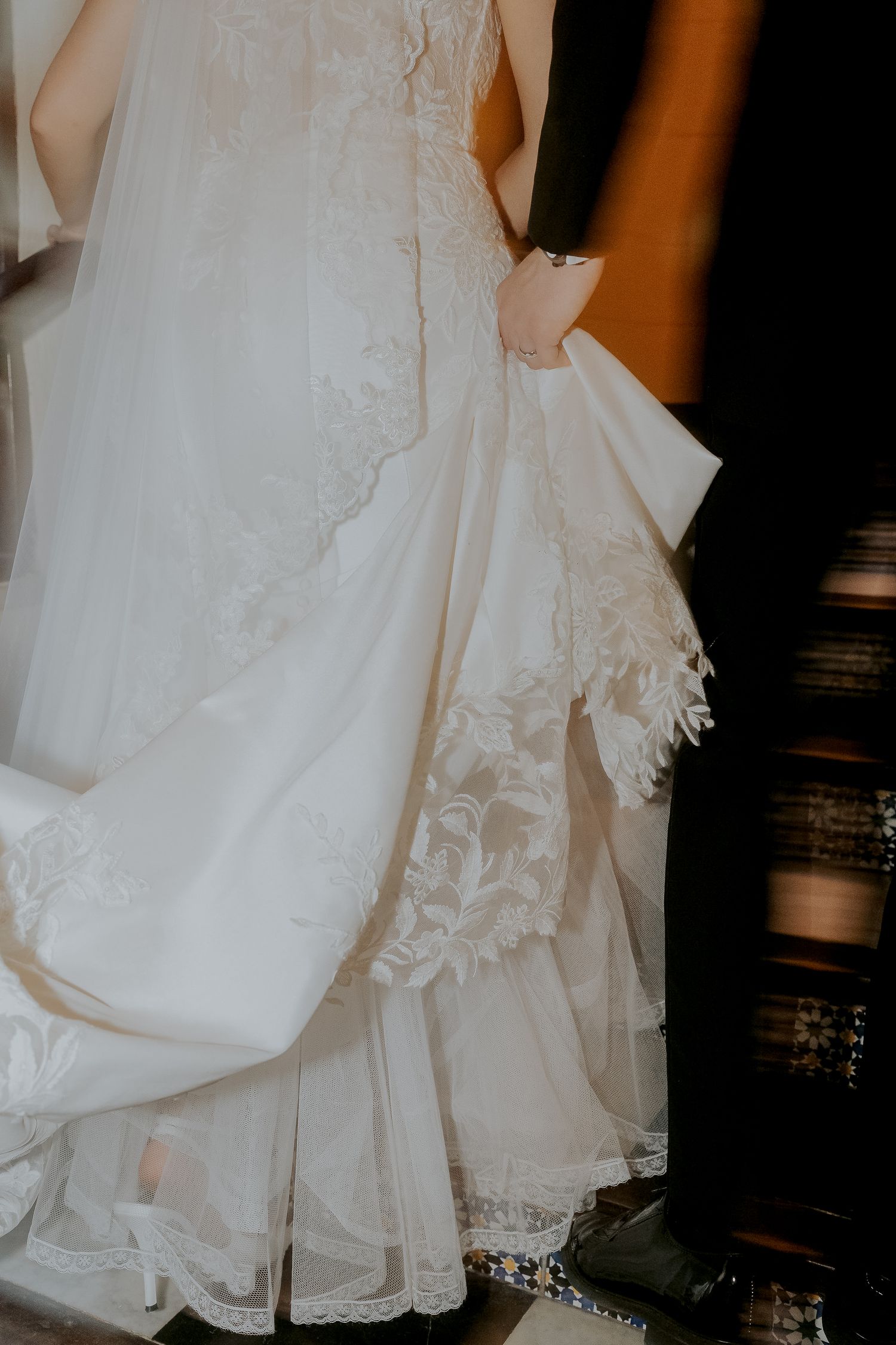 Elegant bride in lace wedding gown with flowing train held by groom in black suit.