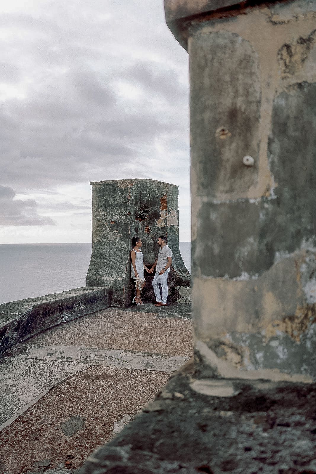 Couple dressed in white sharing a tender moment beside an ancient stone watchtower by the ocean.