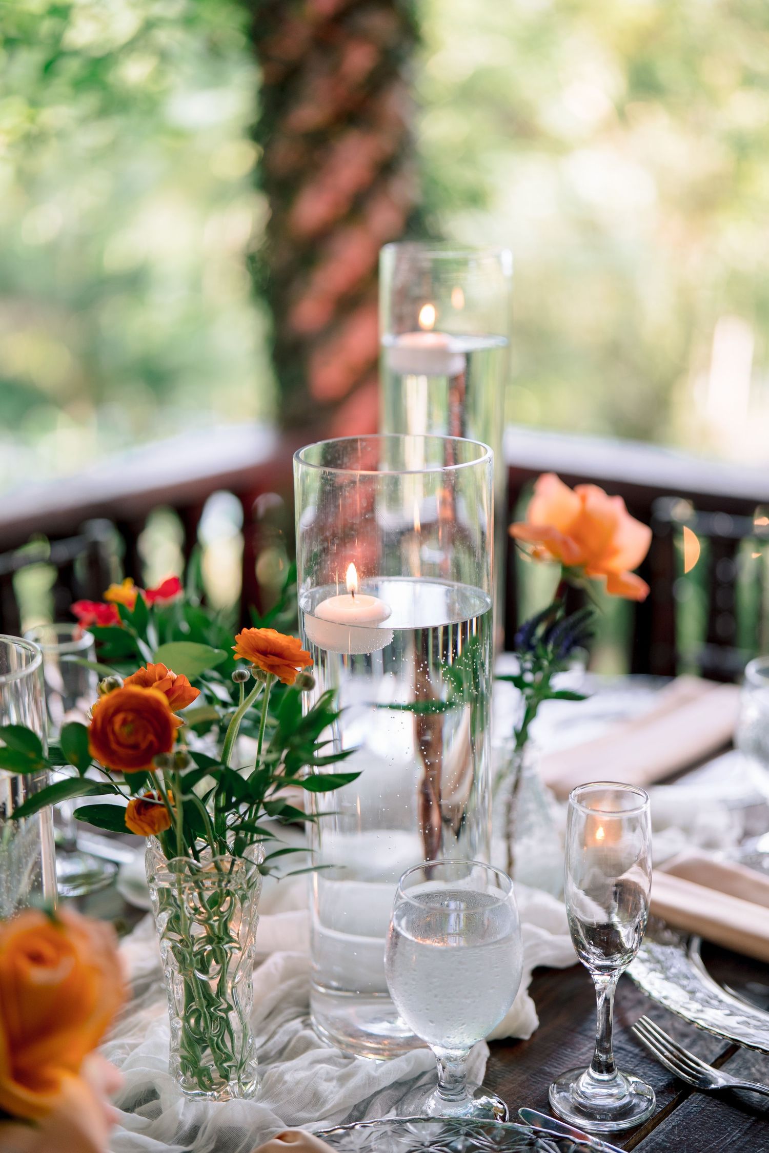 Romantic outdoor wedding table with floating candles in glass cylinders and orange floral arrangements.