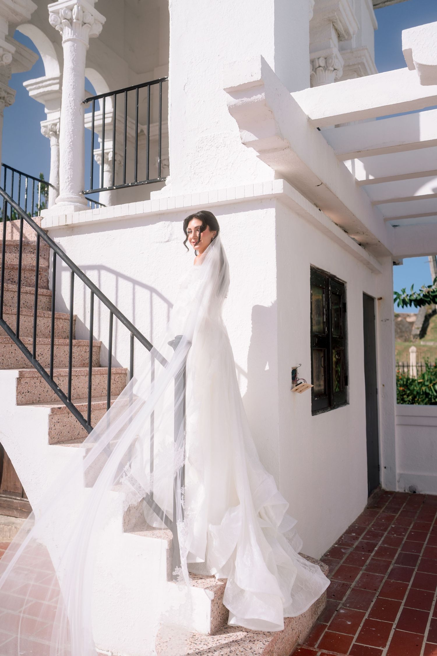 Bride in a flowing white wedding gown and veil stands on outdoor stairs of a white villa.