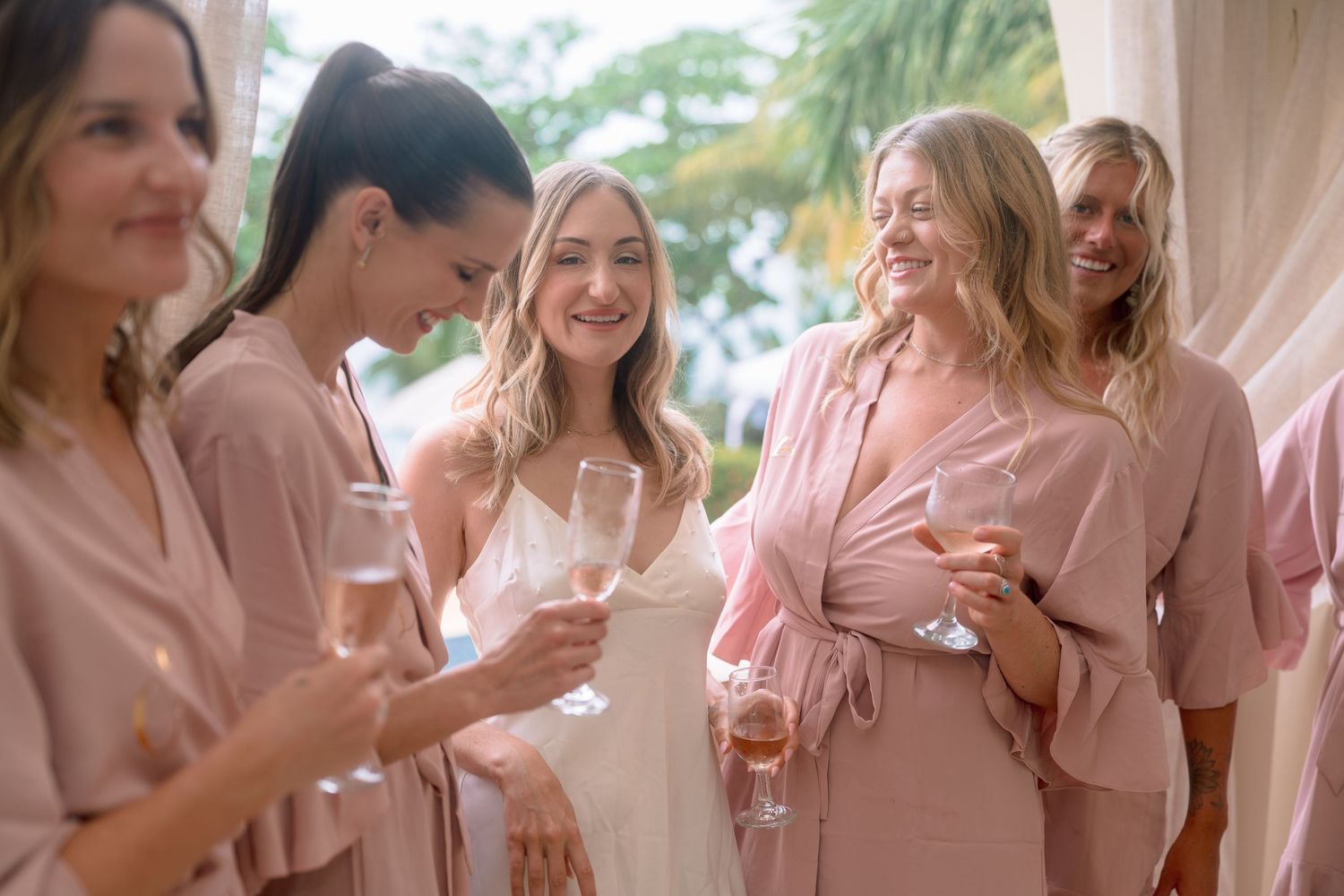 Bridesmaids in pink robes laughing and holding champagne glasses with the bride during wedding preparations.
