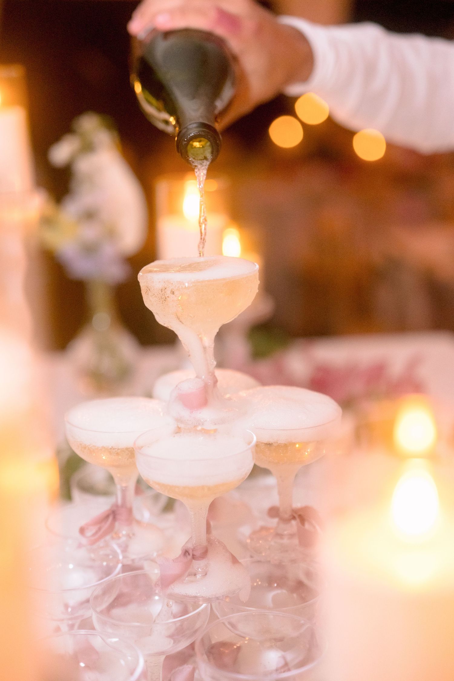 Champagne tower being filled at a wedding reception with warm candlelight glowing in background.