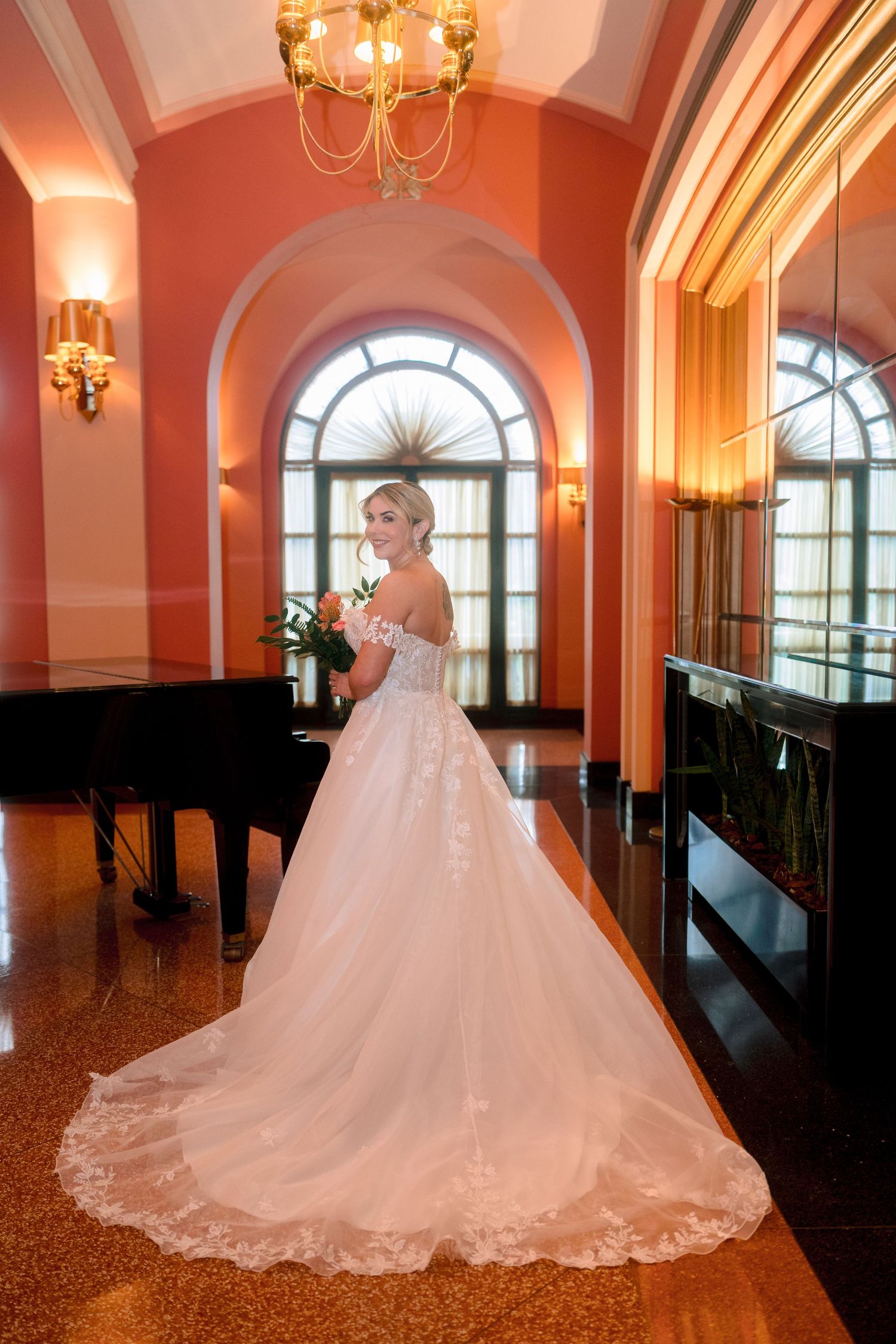 Bride in elegant white off-shoulder ball gown with lace train poses in ornate venue with grand piano.