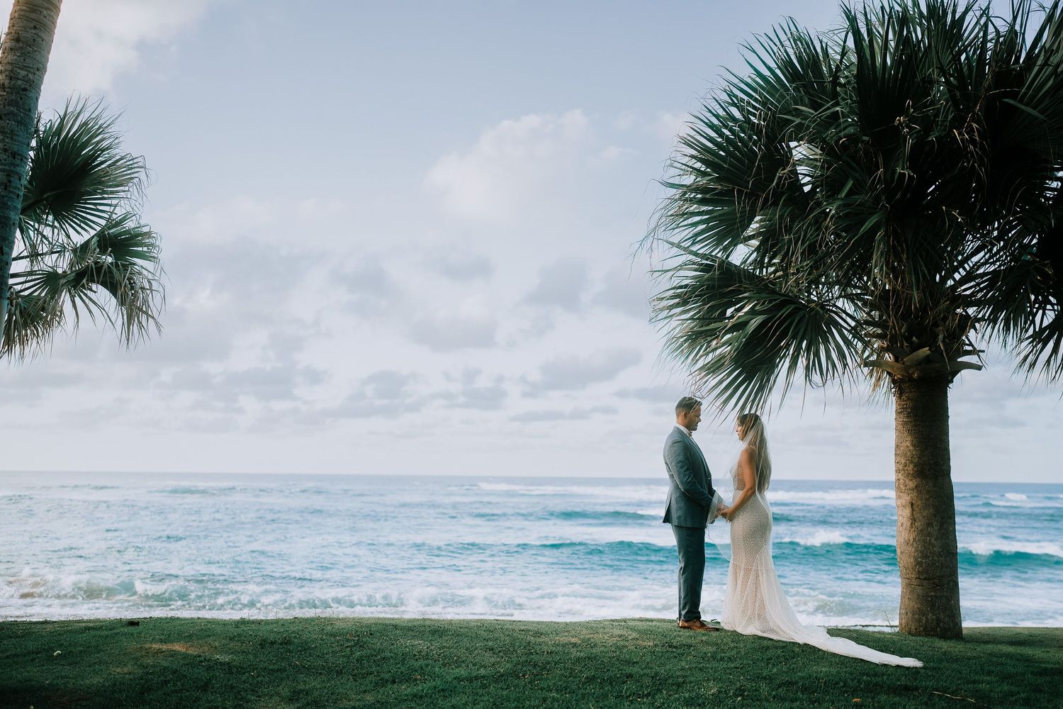 Bride and groom stand hand in hand by a palm tree overlooking a tropical ocean on their wedding day.