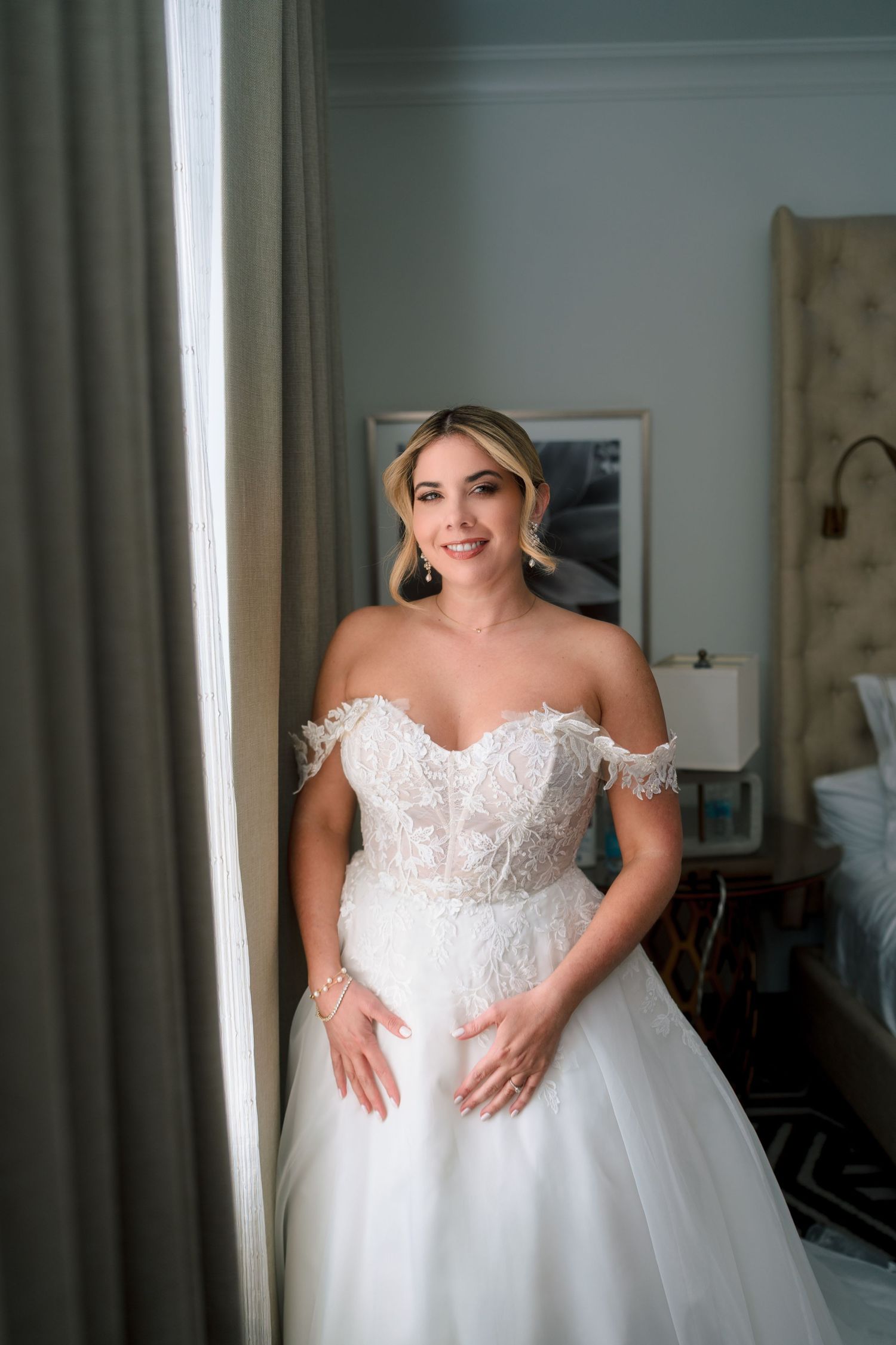 Bride in off-shoulder lace ballgown posing by a window in a softly lit hotel room.
