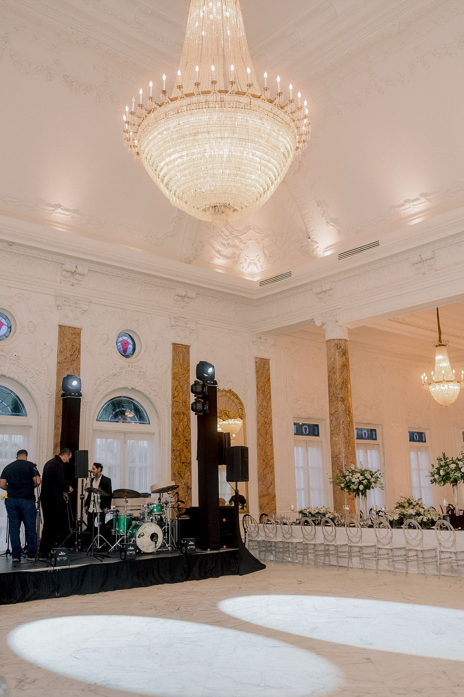 Grand ballroom wedding reception with crystal chandelier, marble floors, and live band stage setup.