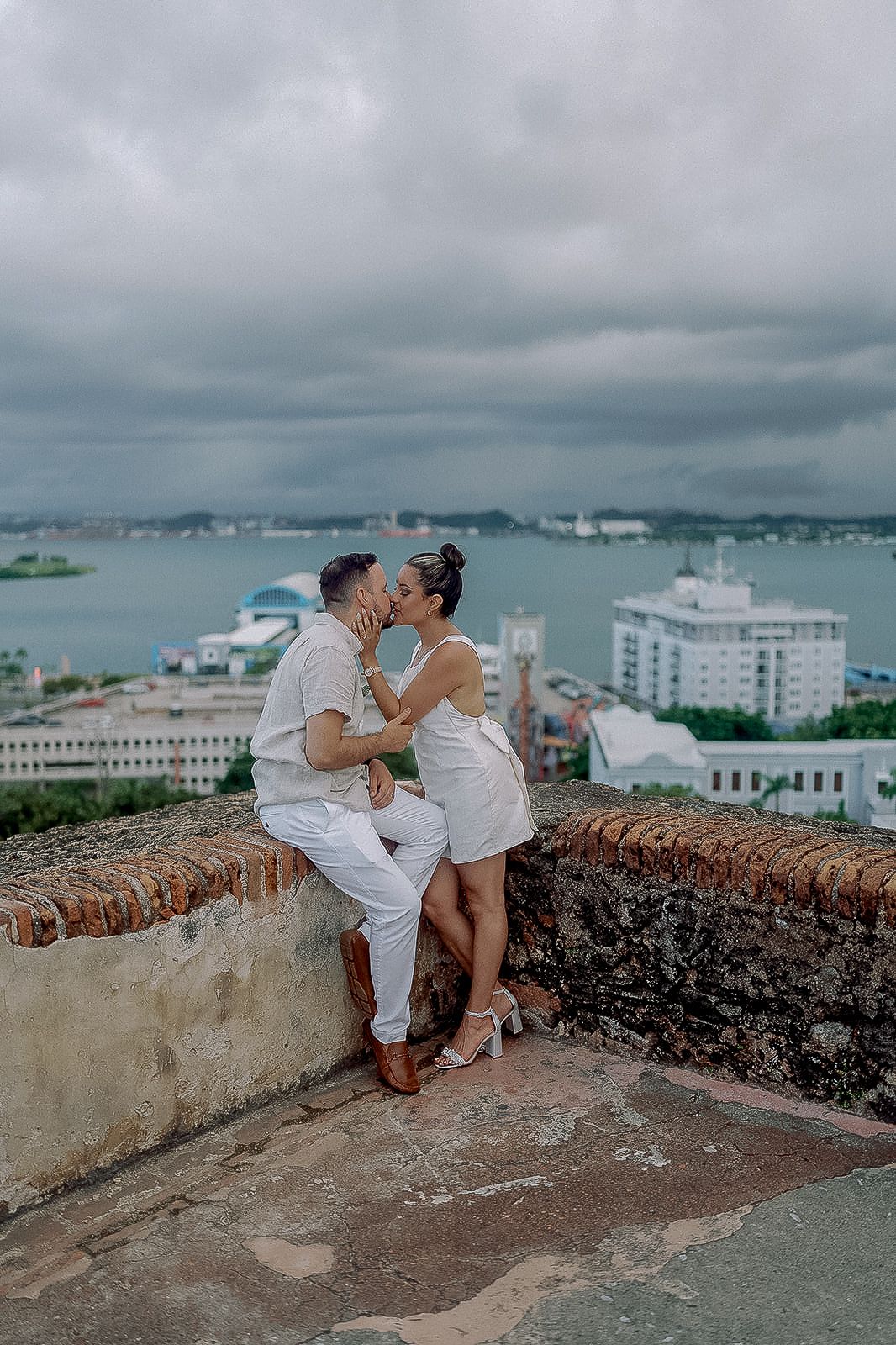 Couple kissing on historic stone wall overlooking San Juan bay in matching white outfits on cloudy day.