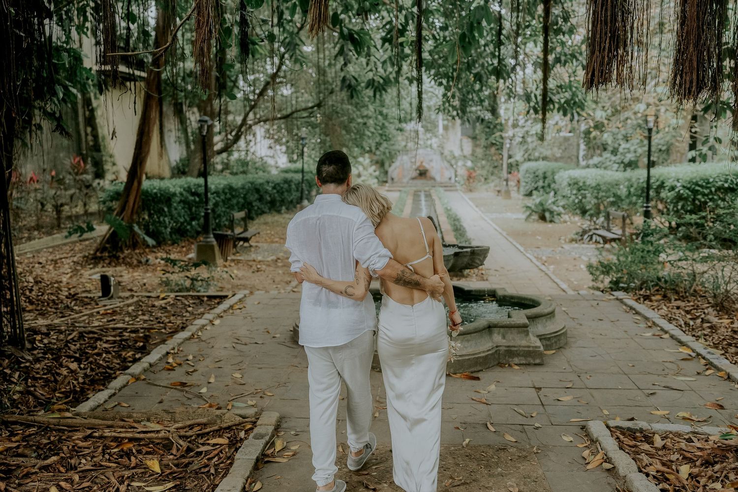 Couple walking hand in hand through a lush garden pathway surrounded by tall trees and greenery.