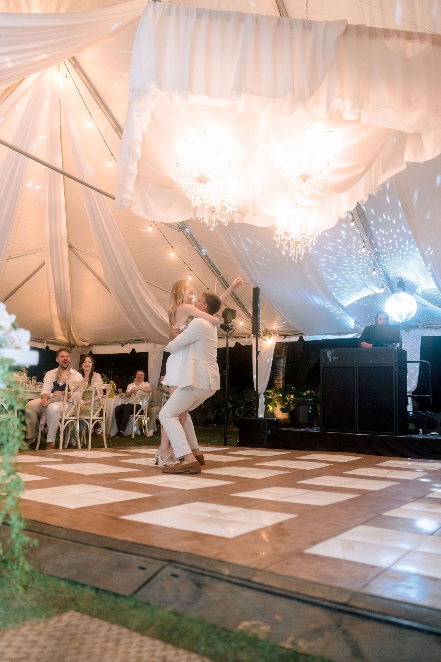 Bride and groom share energetic first dance under elegant chandelier in draped white wedding tent.