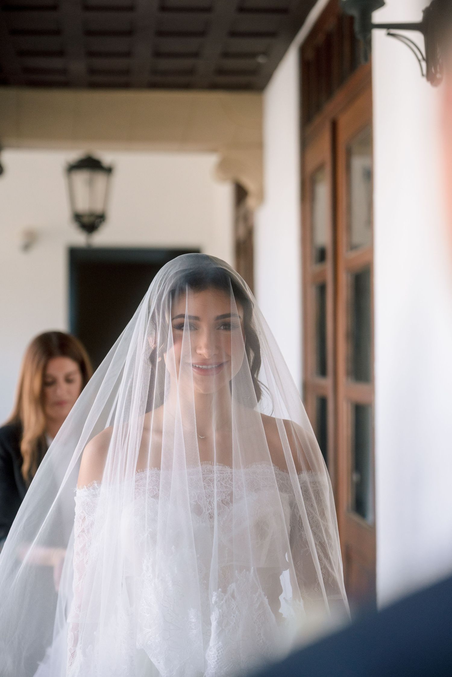 Bride wearing a white lace gown and sheer veil over her face, smiling softly at a elegant venue.