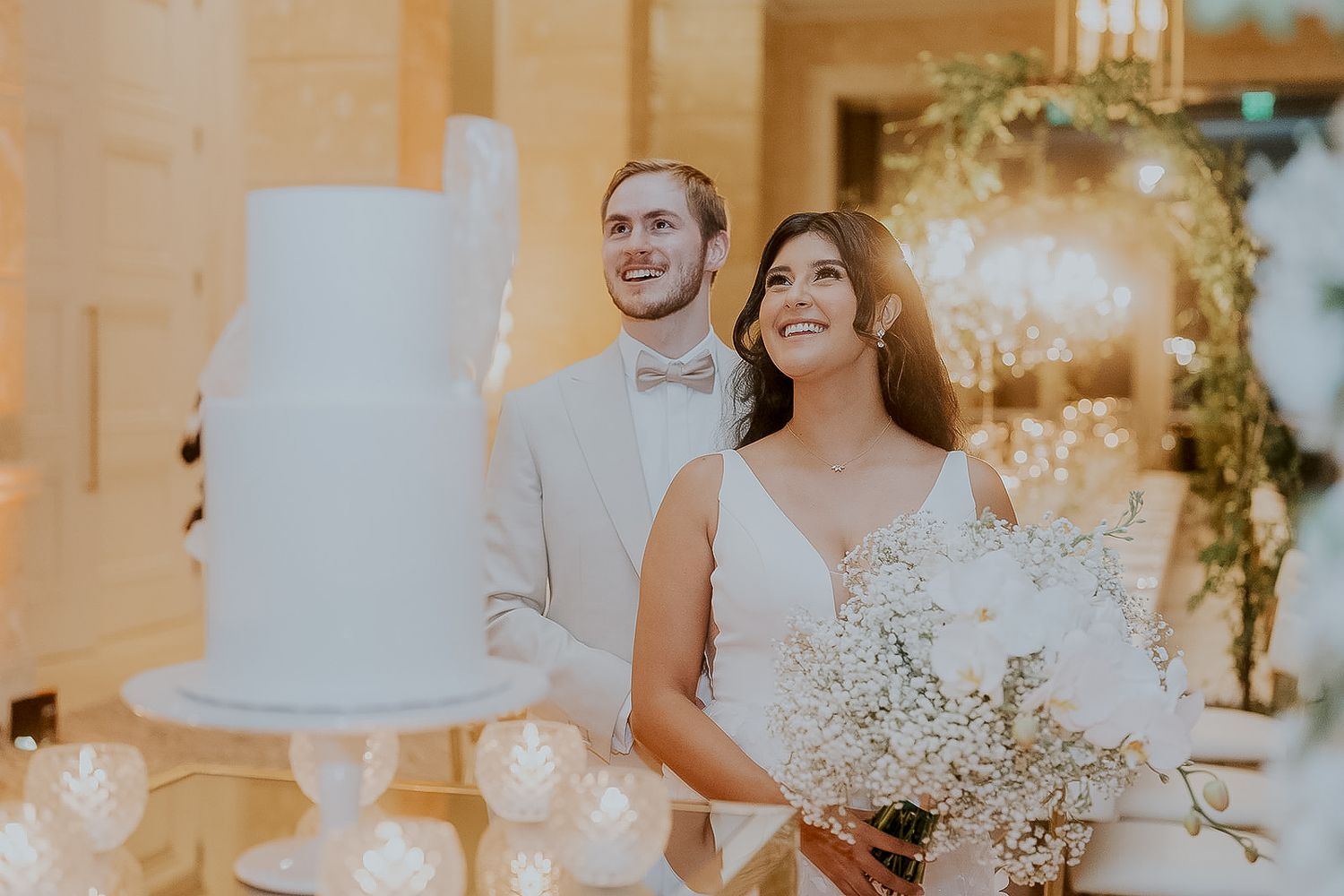 Happy bride and groom stand beside a white tiered wedding cake at a warmly lit reception.