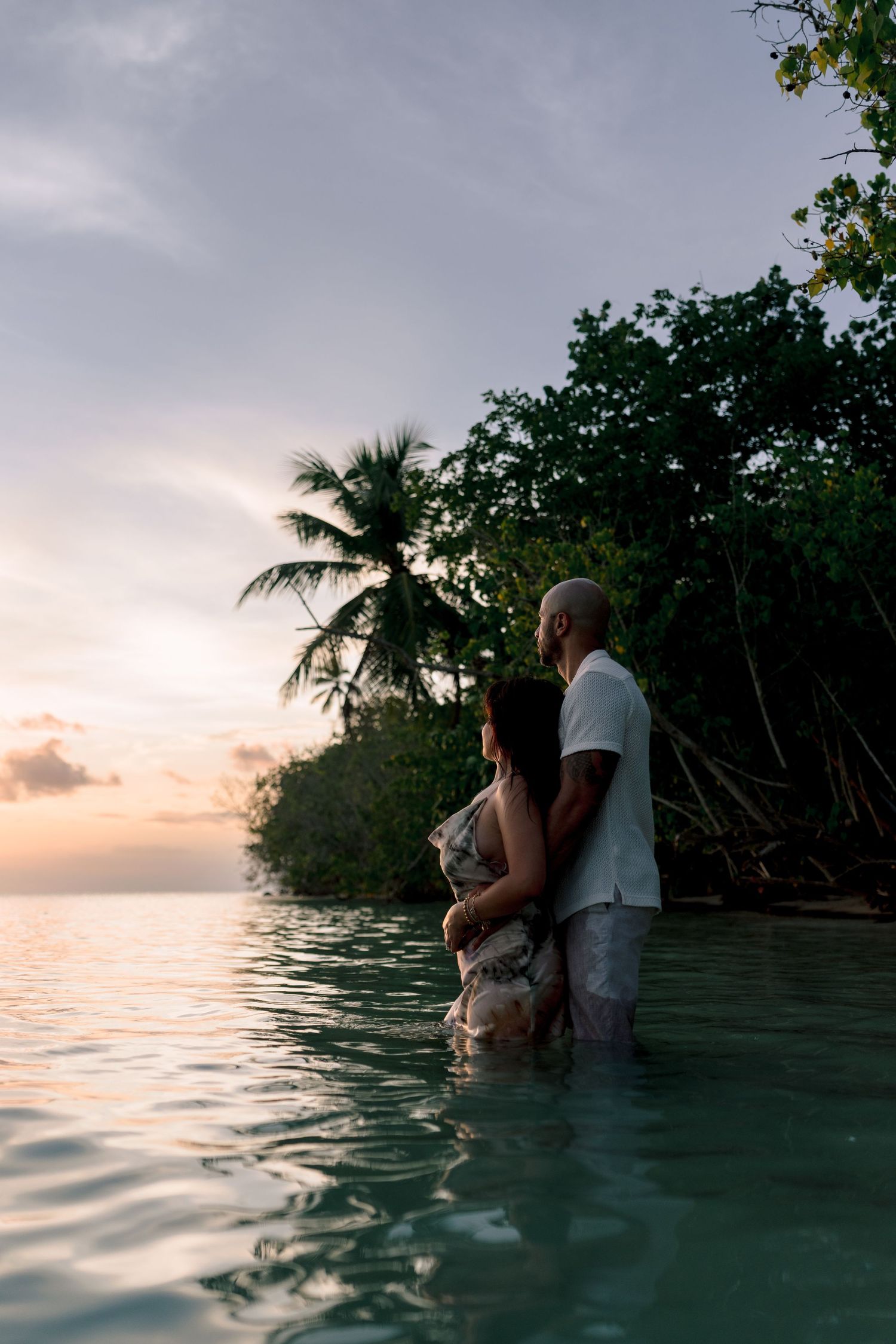 Couple standing in turquoise water at sunset near tropical palm trees, sharing a romantic embrace.