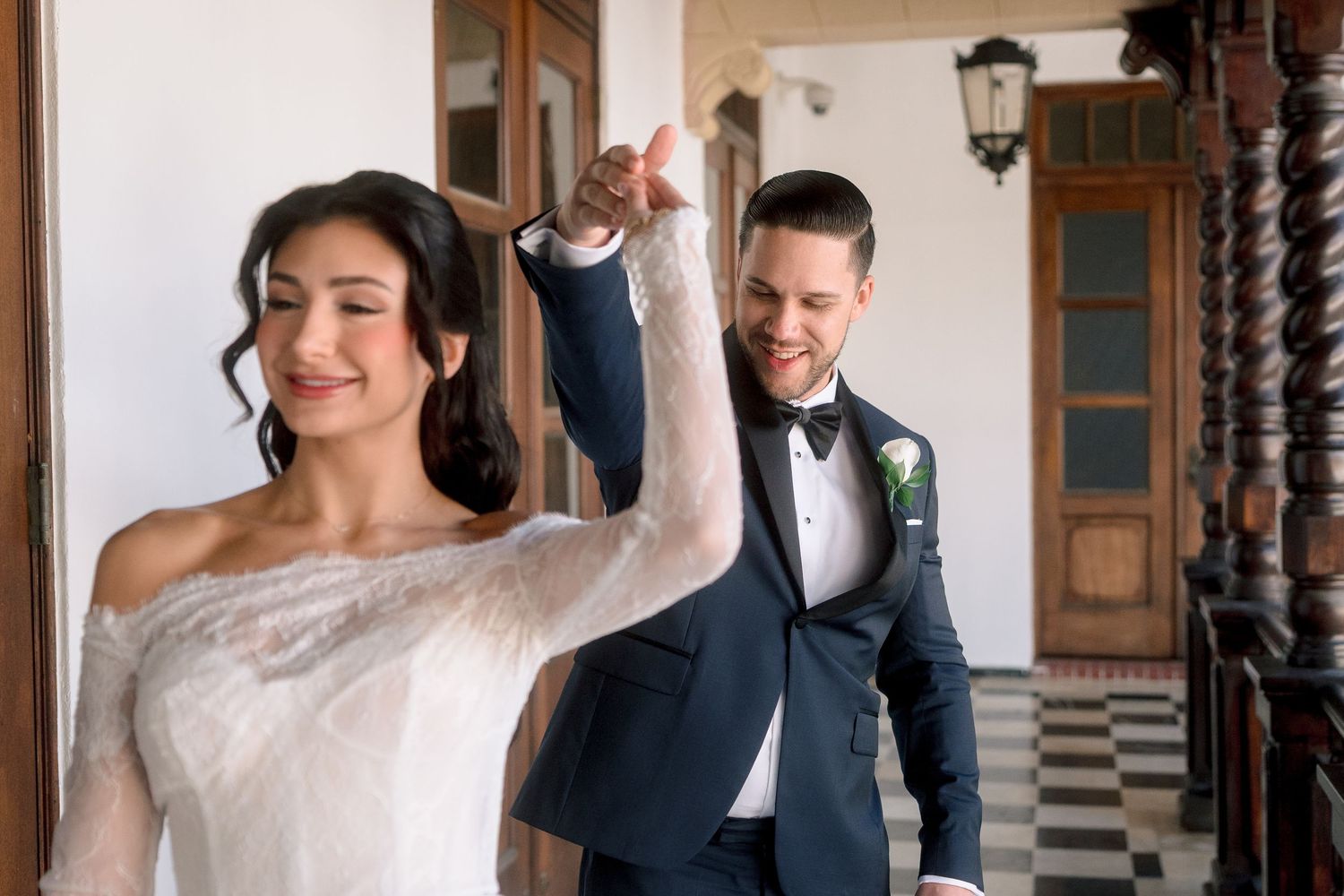 Groom in navy tuxedo twirling bride in elegant white lace off-shoulder wedding gown indoors.