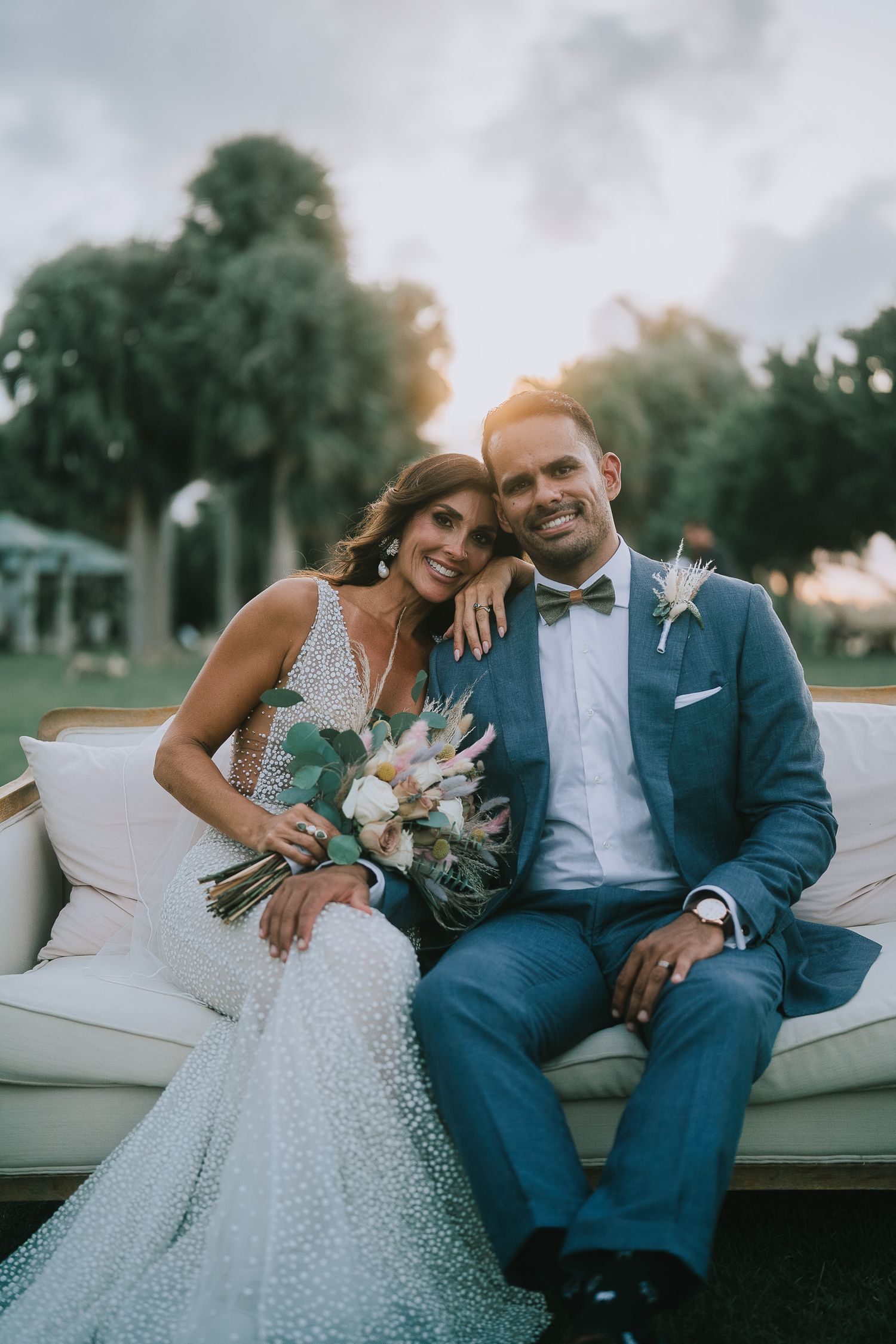 Bride and groom sitting on white outdoor sofa at sunset, she holds a bouquet, he wears a blue suit with bow tie.