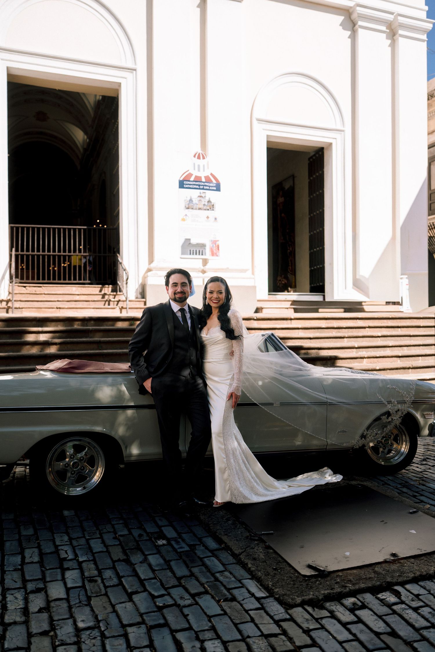Bride in fitted white gown with veil and groom pose beside vintage convertible on cobblestones.