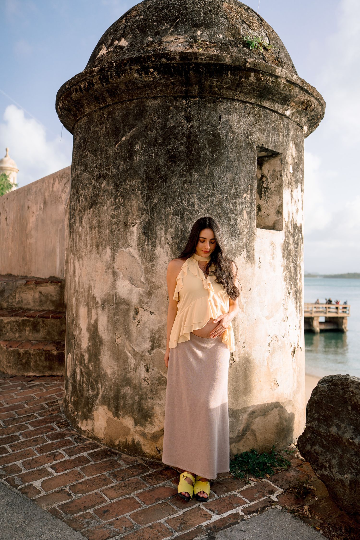 Woman in peach top and maxi skirt poses by ancient stone fort tower near waterfront.
