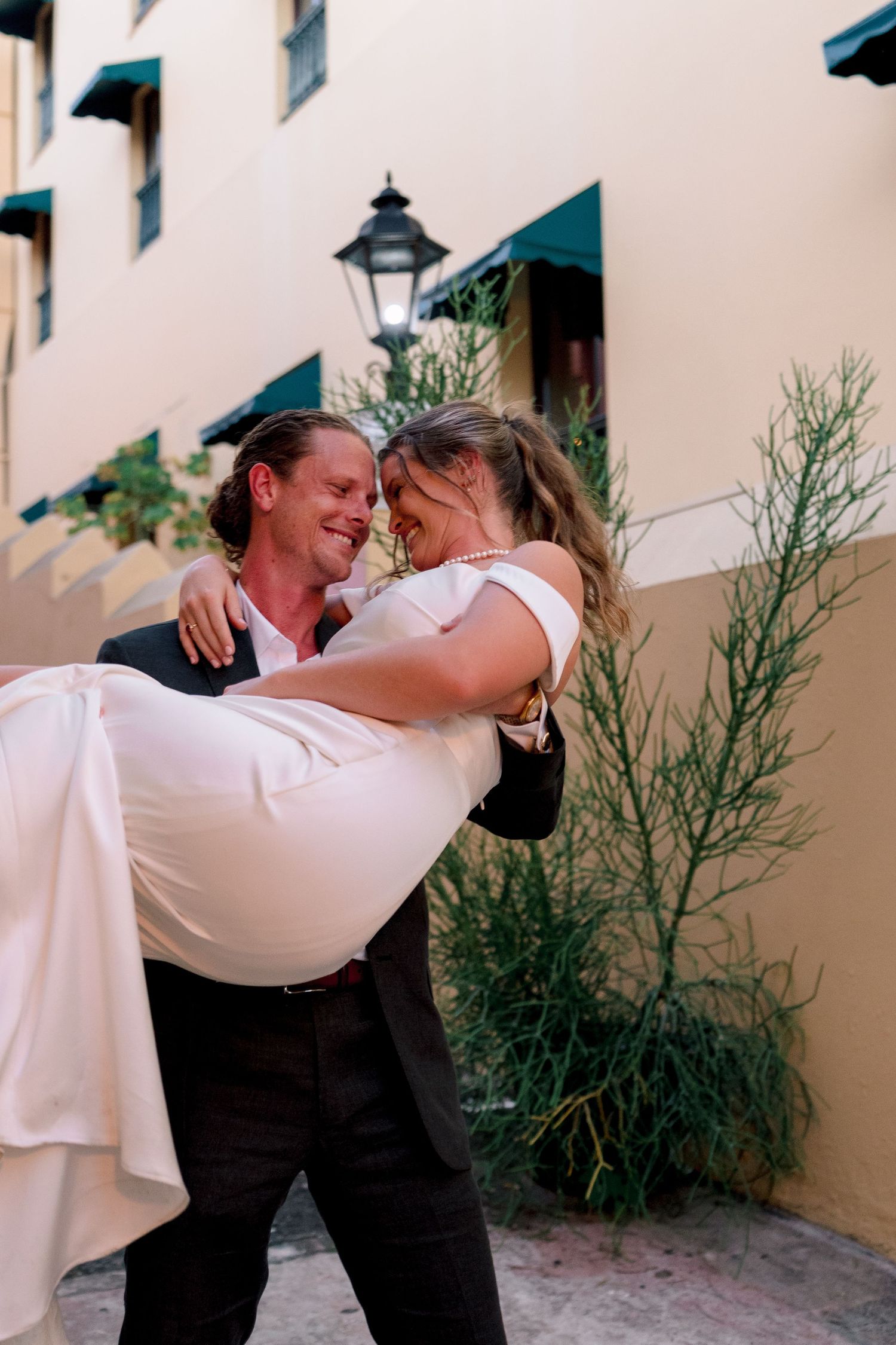 Groom carrying bride in white dress outside a yellow building with green awnings on their wedding day.